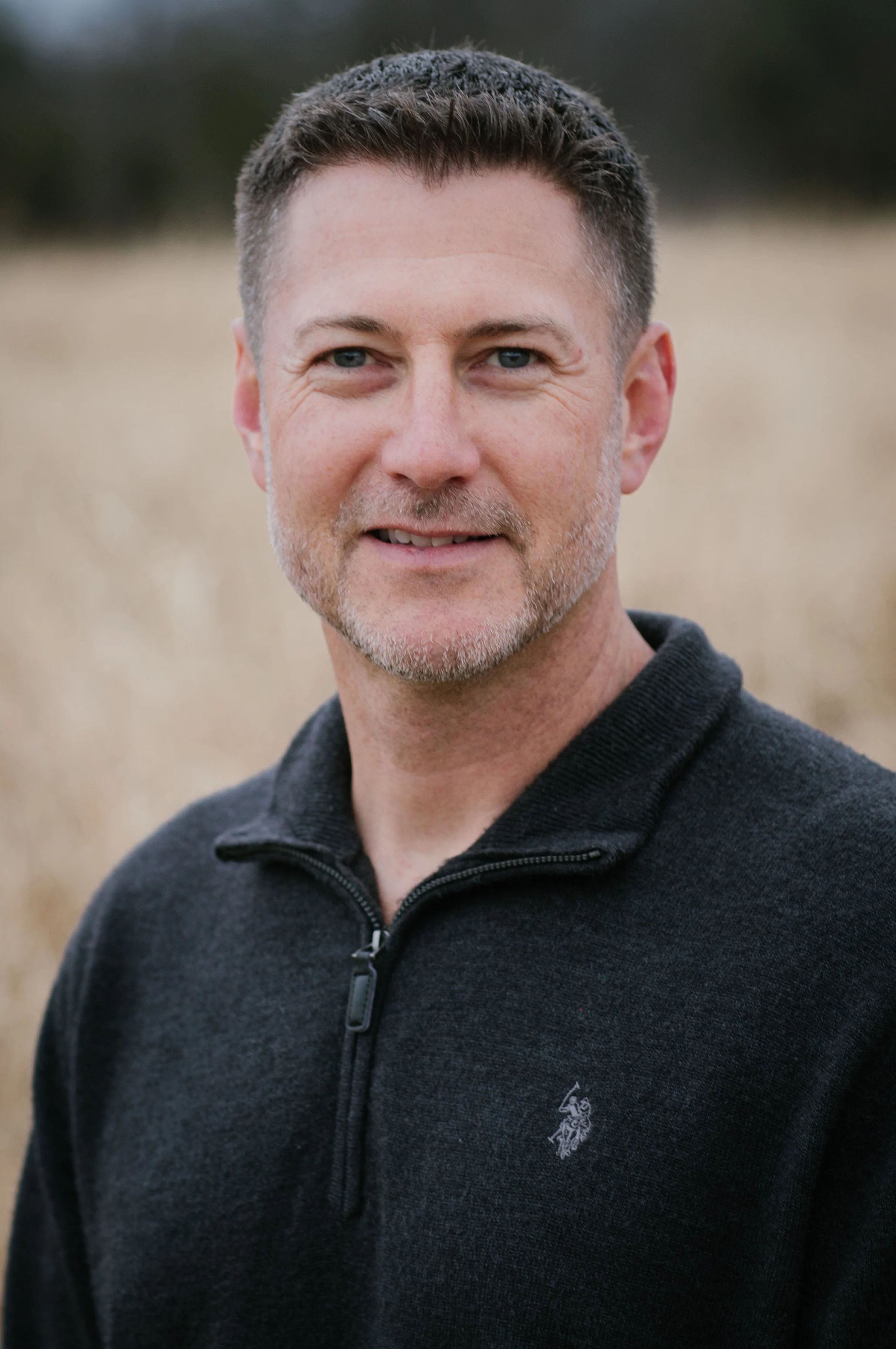 A man with short brown hair, a light beard, and blue eyes, wearing a black zip-up sweater with a small logo, standing outdoors in a field with a blurred background.