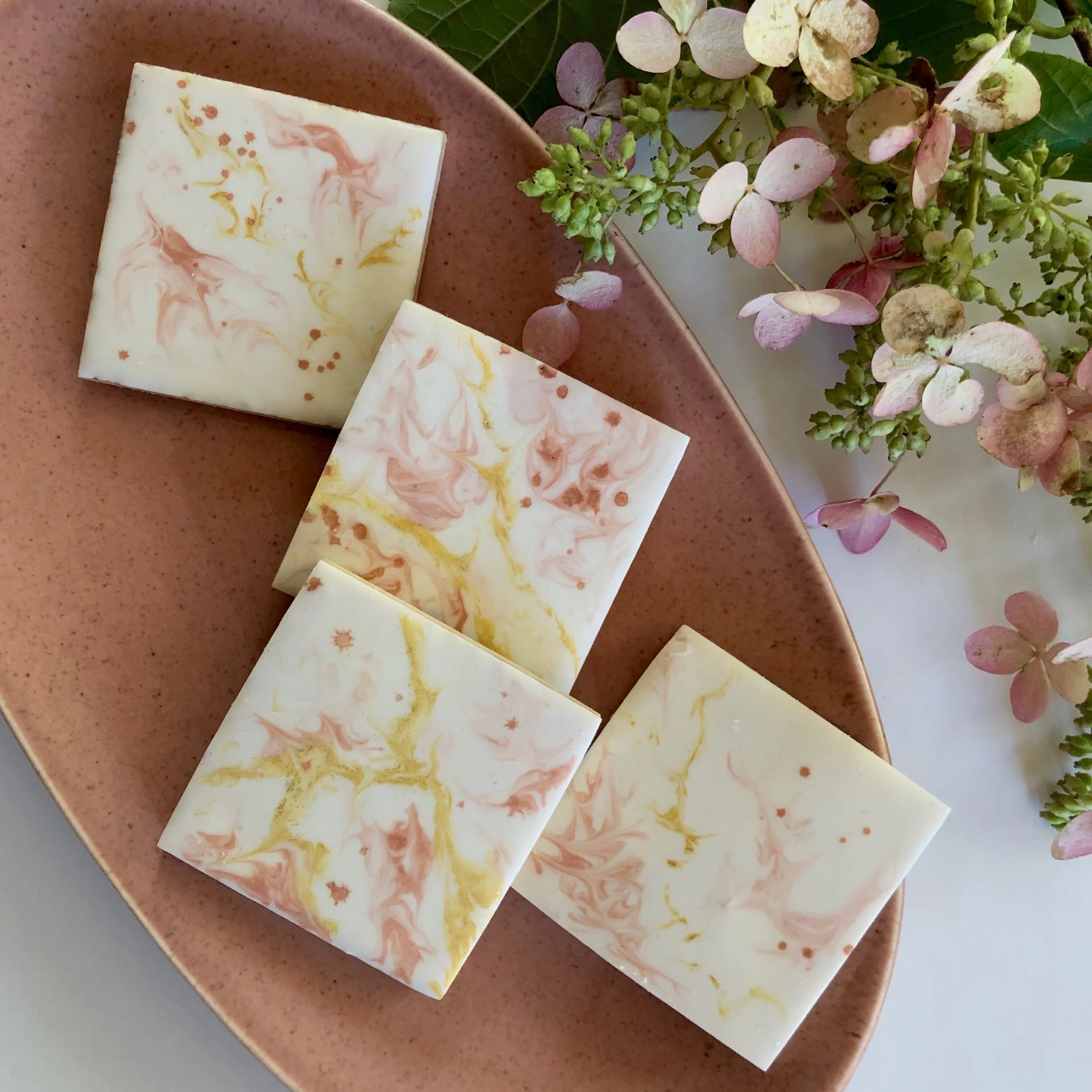 Marbled soap bars with pink, yellow, and white swirls, placed on a pink plate, with pink and green hydrangea flowers nearby.