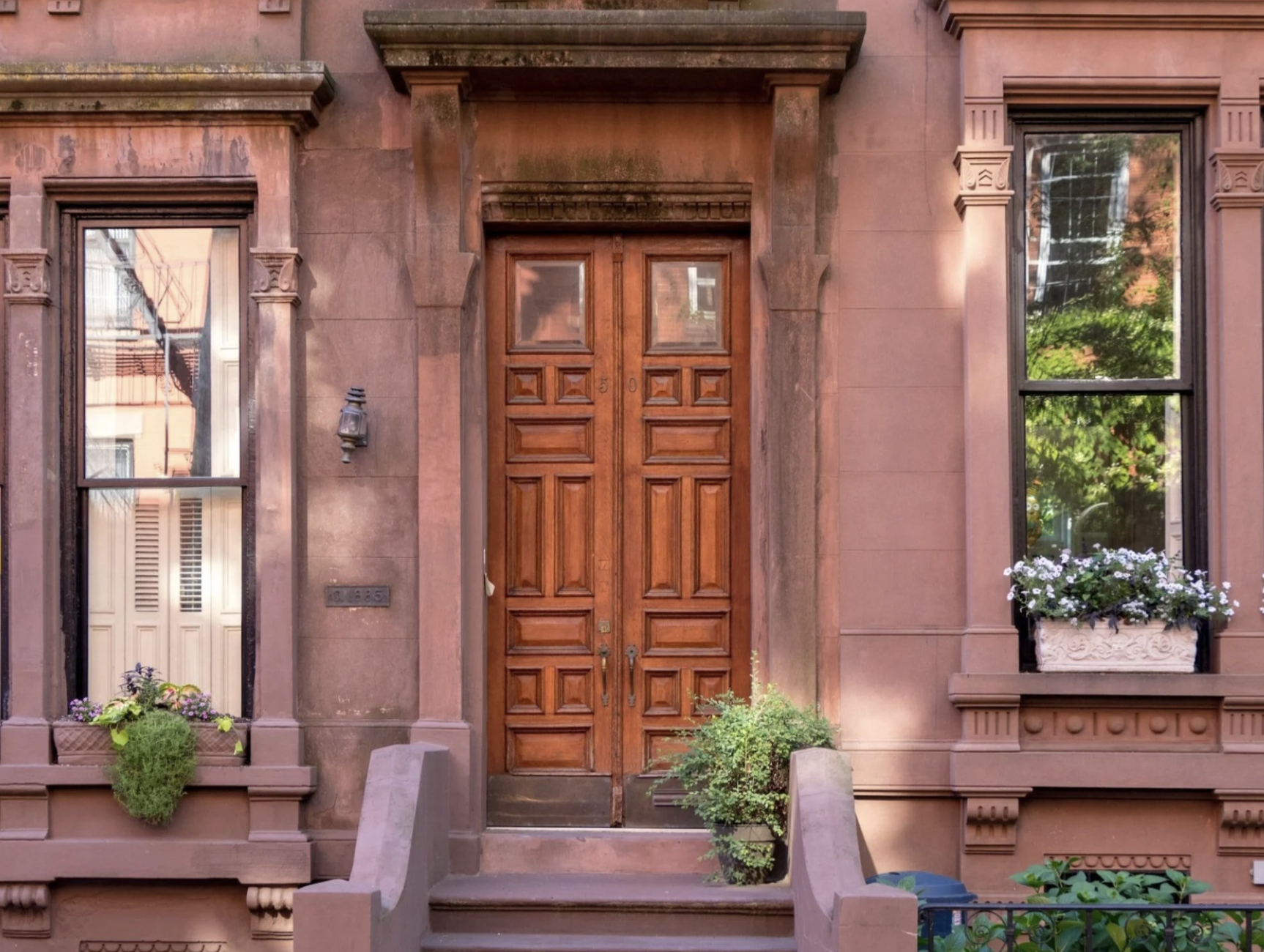 Brownstone building facade with wooden front door, two large windows with black trim, flower boxes, and potted plants.