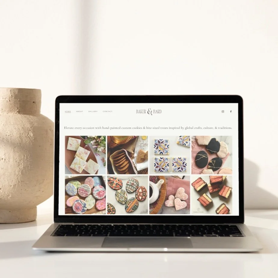 A laptop on a white desk displaying a website for Baker & Bard, featuring various images of handcrafted cookies and treats, with a beige textured vase beside it.