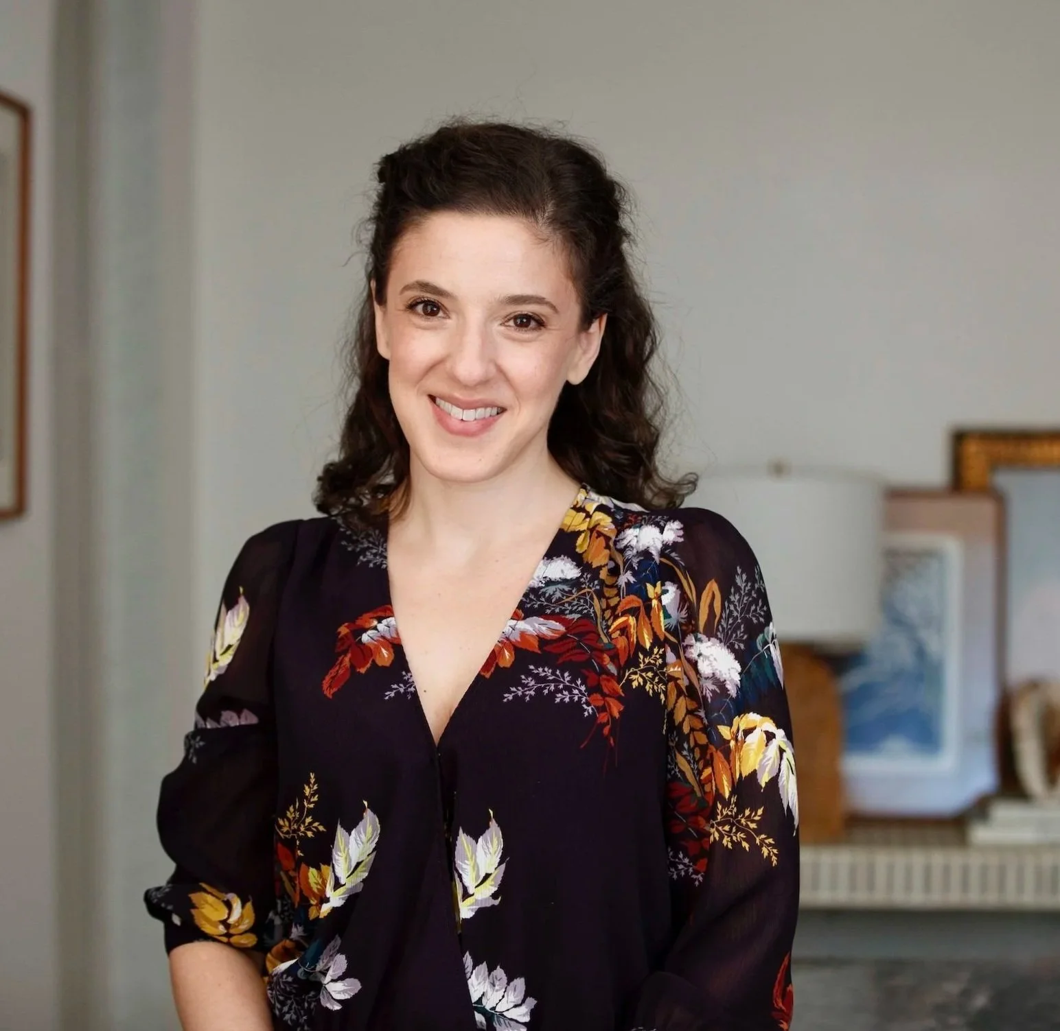 A woman with dark, curly hair smiling, wearing a black floral dress, standing in a room with light-colored walls and furniture in the background.