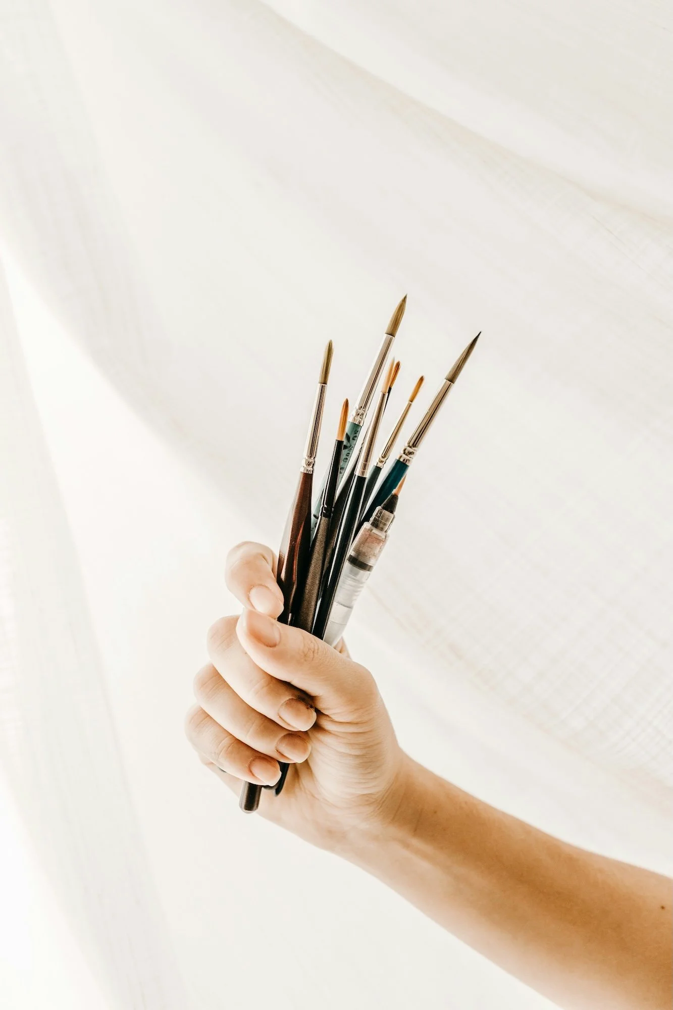 Hand holding a bunch of watercolor paintbrushes with metallic handles and different colored bristles.