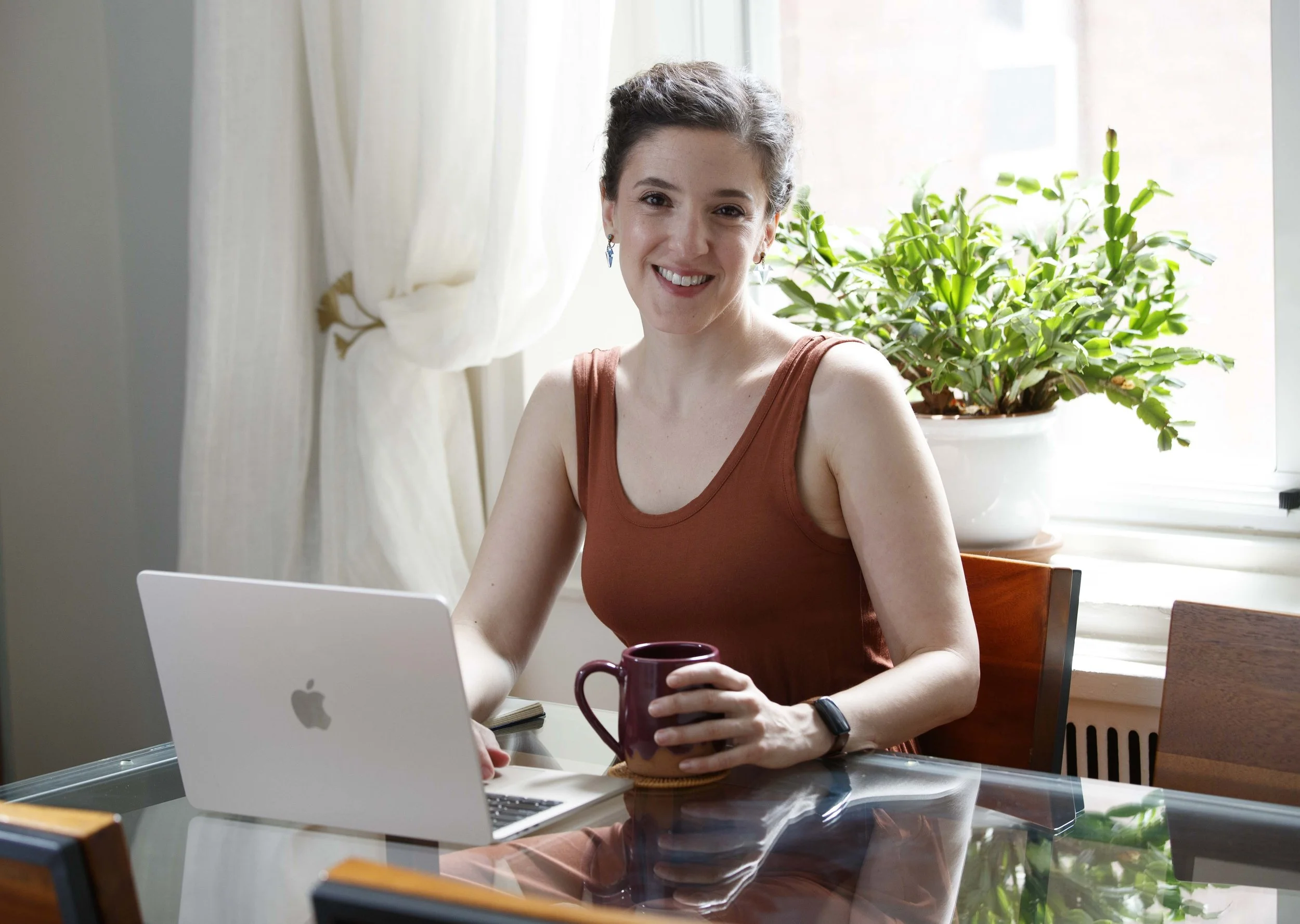 A woman sitting at a glass table with a silver MacBook laptop in front of her, holding a maroon mug, with a green potted plant and cream-colored curtains behind her, smiling at the camera.