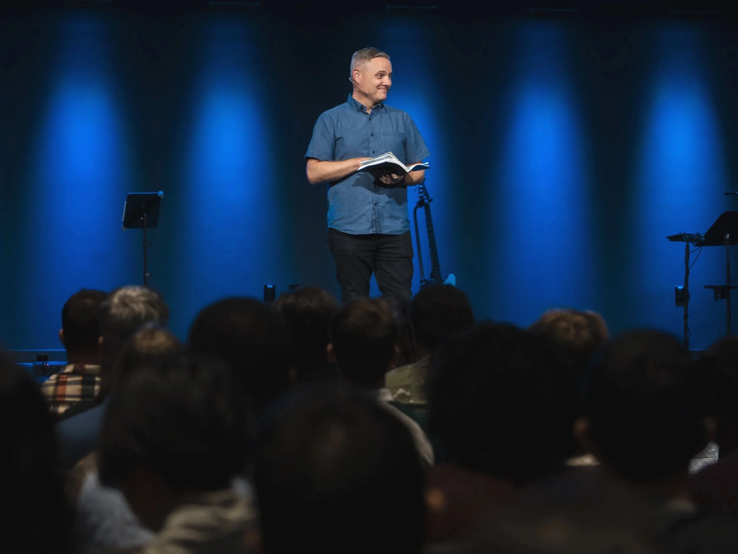 A man wearing a blue shirt stands on a stage holding a book, with a dark background and audience in front.