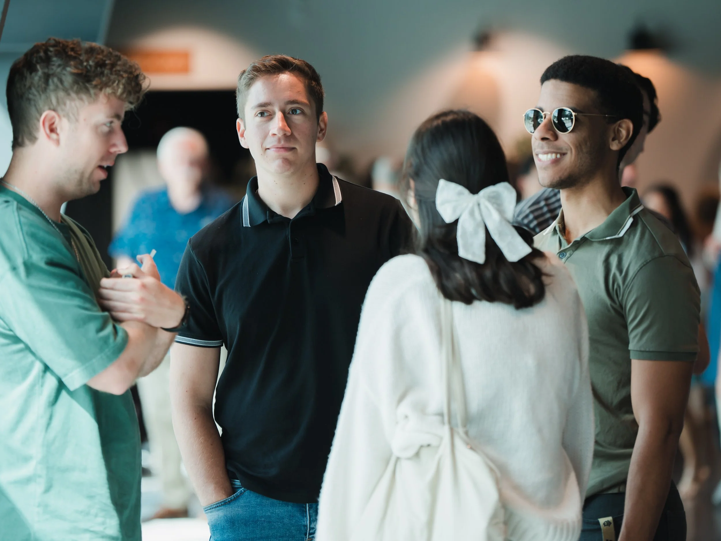 A group of four young people engaged in conversation, three men and one woman, in an indoor setting with blurred background and warm lighting.