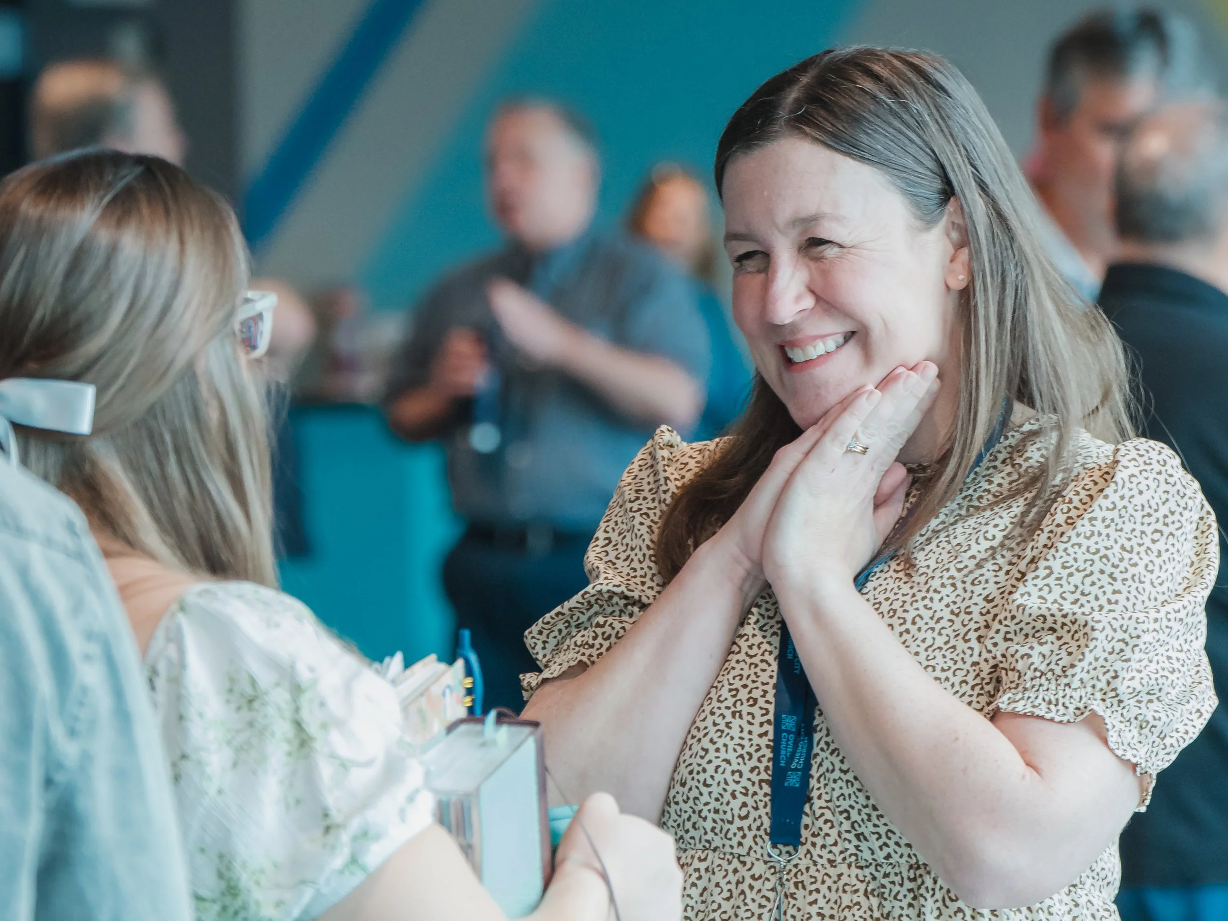 A woman smiling and touching her face with her hands in a crowded indoor setting, at a church service listening with love.