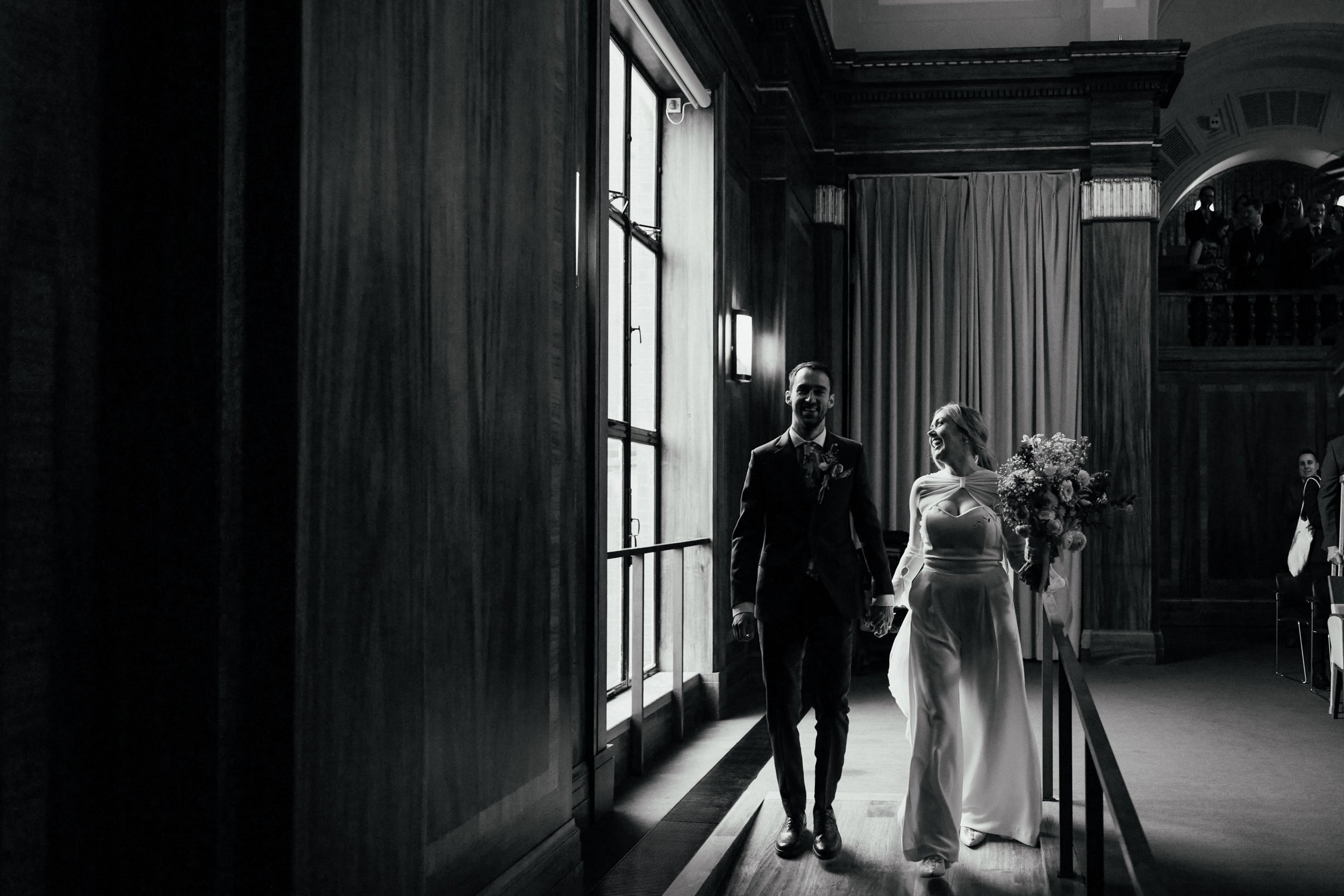 A black and white image of a just married couple joyfully walking out of their ceremony at Stoke Newton Town Hall, they are lit dramatically by window light from the left hand side. The bride is mid stride and we can see that she is wearing a wide l