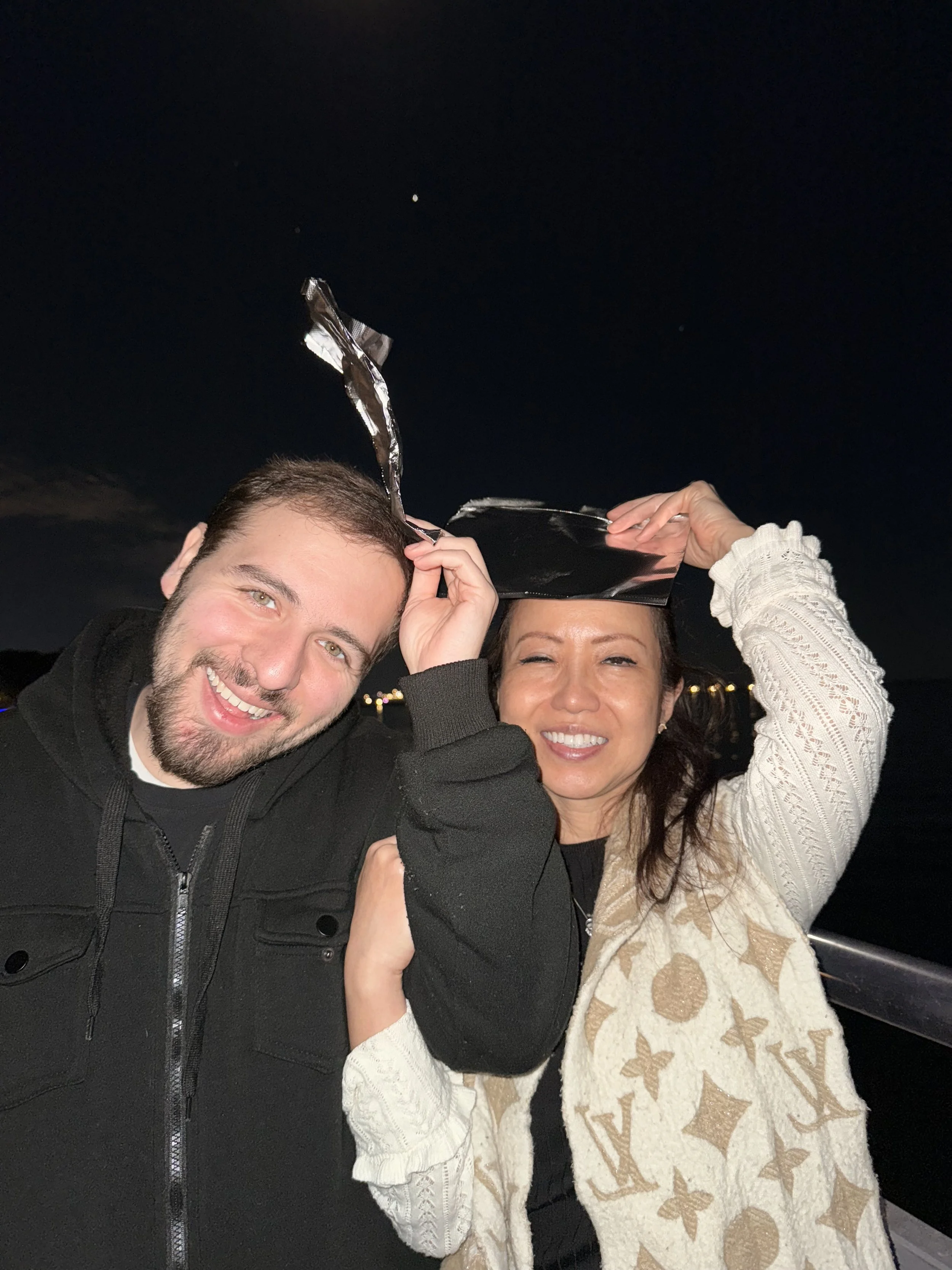 Two people smiling at night: one is a man with a beard, wearing a black jacket; the other is a woman with long dark hair, wearing a beige patterned jacket, holding a shiny tin-foil hat above her head.