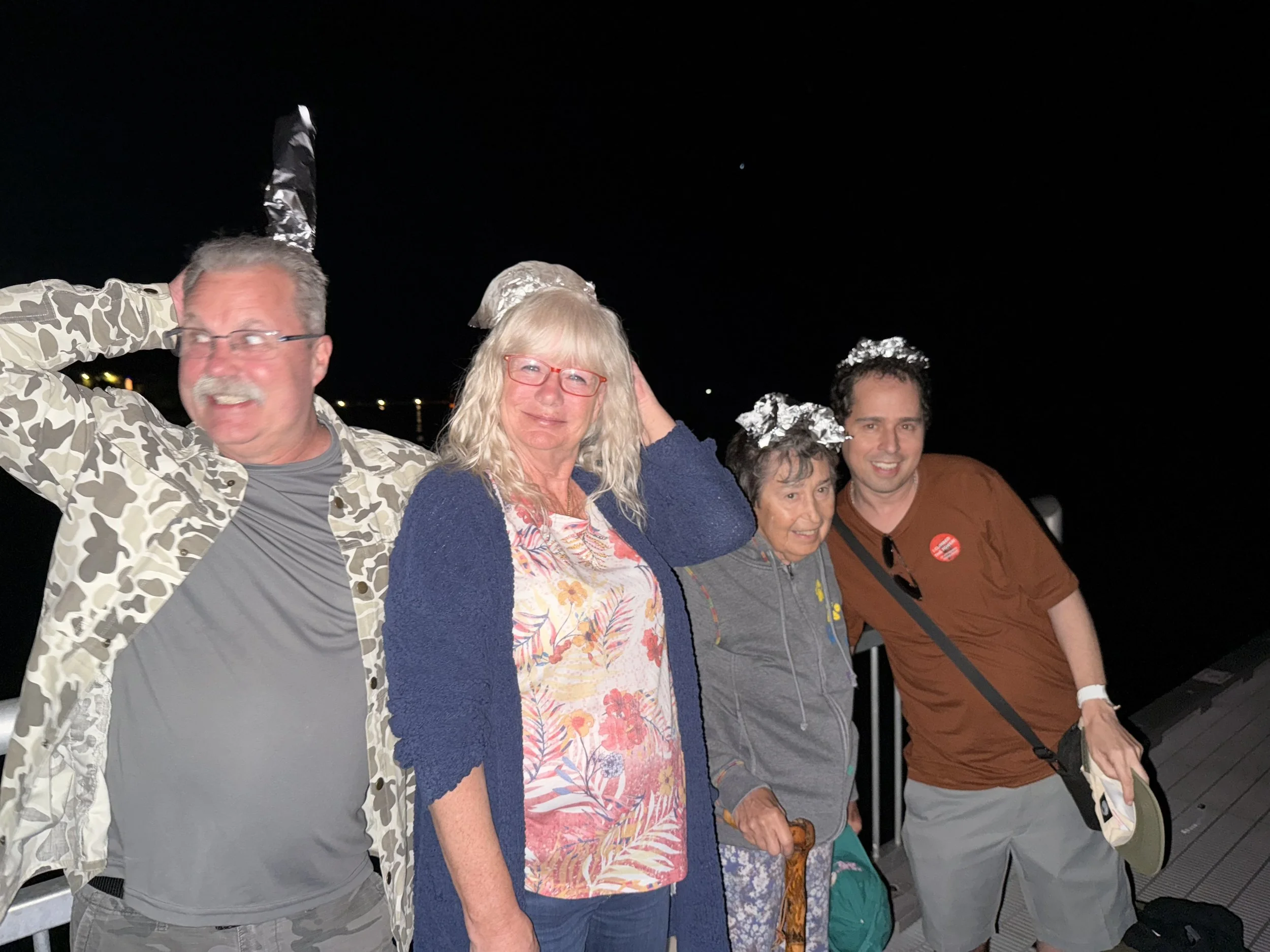 Four people standing outdoors at night, celebrating with aluminum foil hats, smiling, with a dark sky and distant lights behind them.