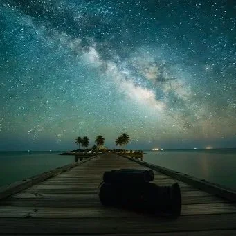 Night sky with stars and the Milky Way over a wooden pier extending into a body of water in Key West, Florida, with palm trees in the distance.