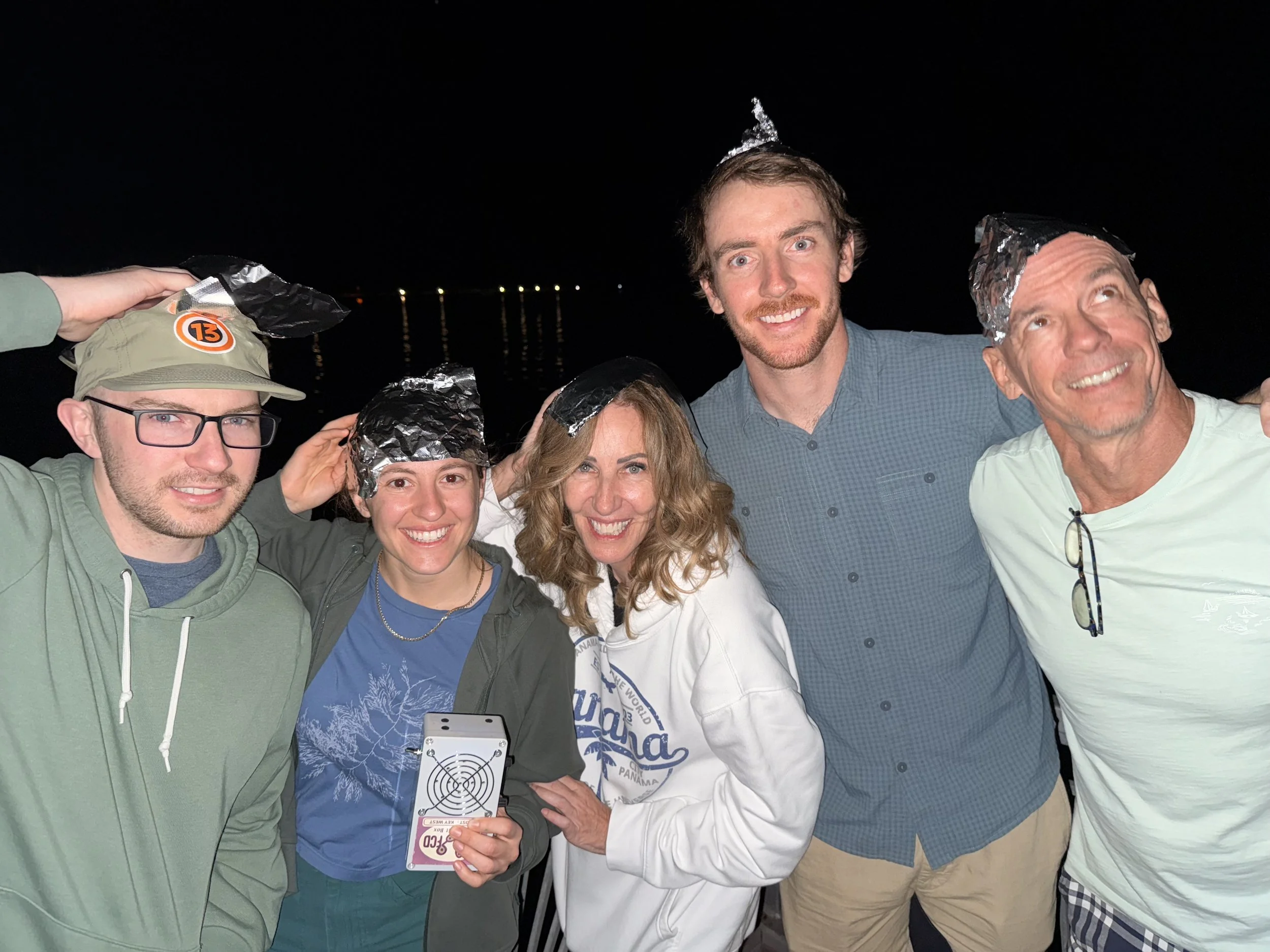 Group of five people at night, some wearing tin foil hats, smiling and posing for a photo, with a dark sky and water in the background.