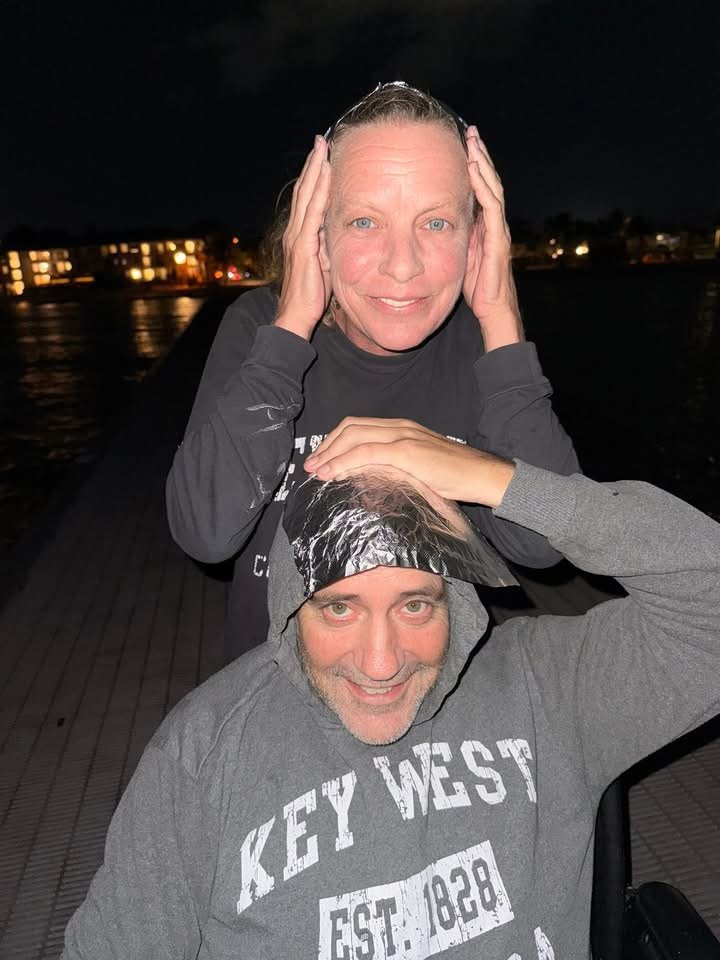 Two adults, a woman and a man, posing playfully outdoors at night. The woman is leaning over the man, holding her hands on her head, and smiling at the camera. The man is wearing a hoodie with a shiny cap, smiling, and touching his head. The background shows a dark sky and some lights from buildings across a body of water.
