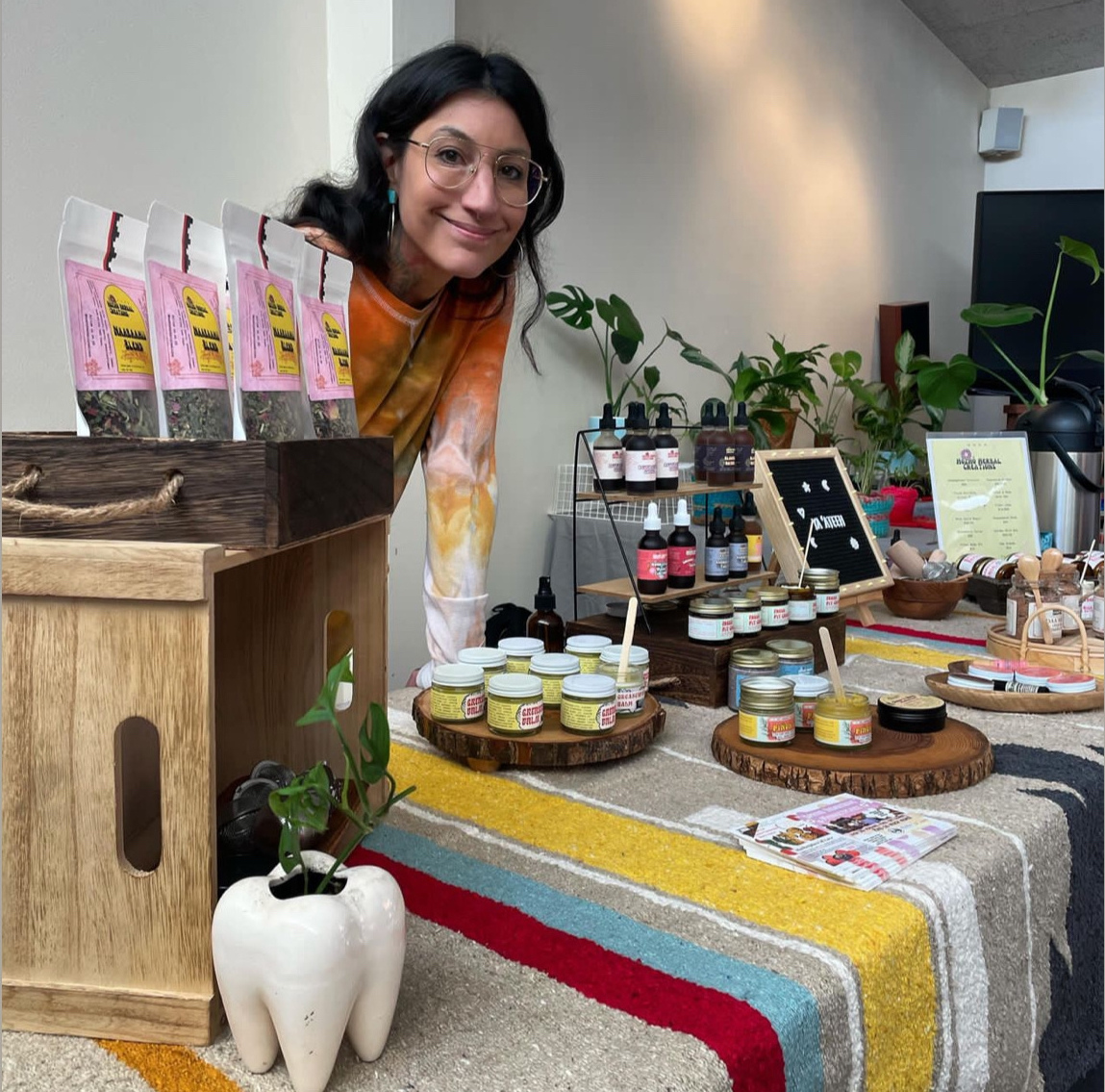 A woman smiling at a market stall displaying various herbal products, including jars, bottles, and herbal blends, with plants and a colorful striped tablecloth in the background.