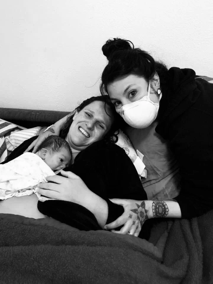 A woman lying in bed holding a newborn baby, with a woman wearing a face mask leaning over her, all appearing happy and close.