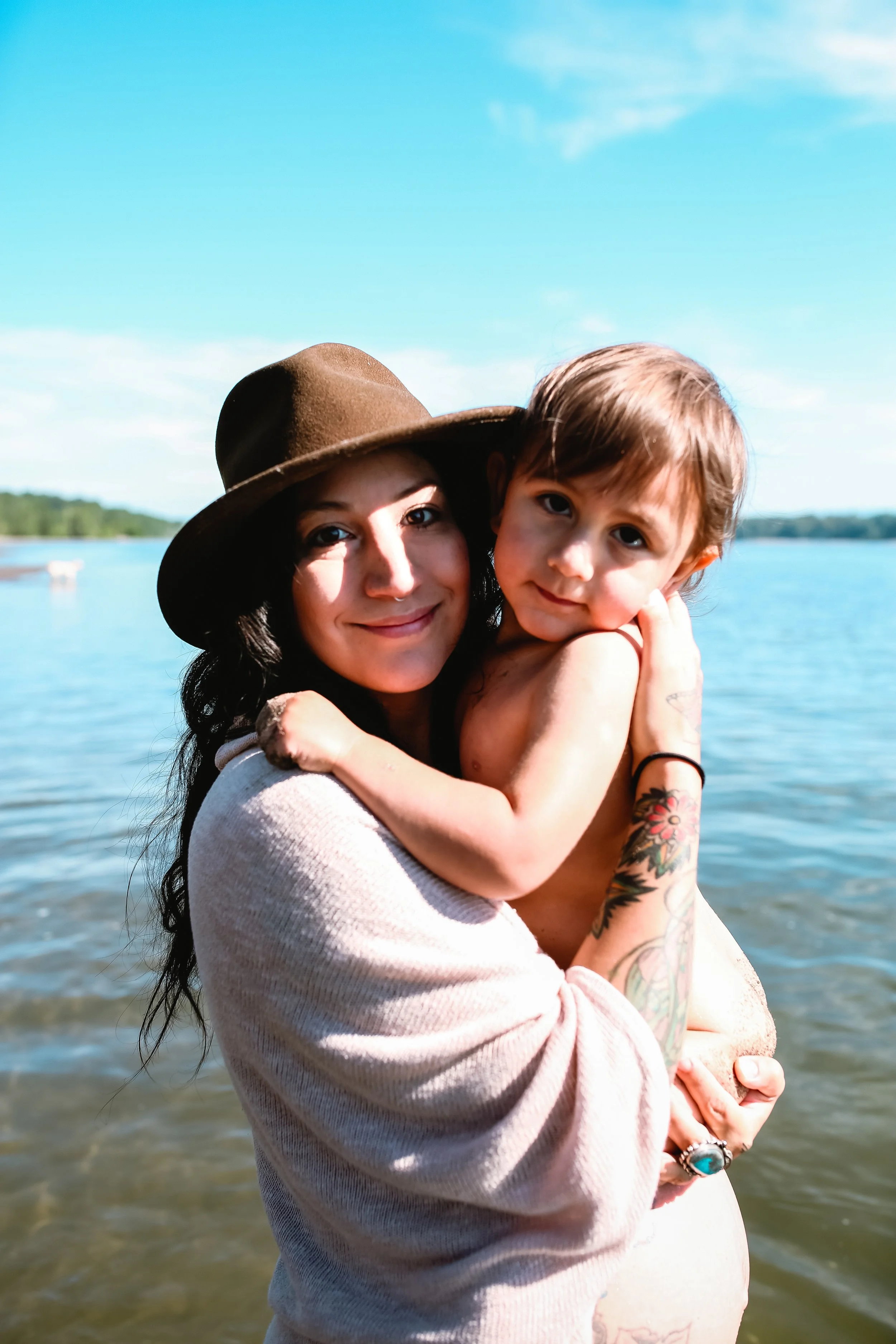 A woman with tattoos and a hat holding a young child by the water on a sunny day.