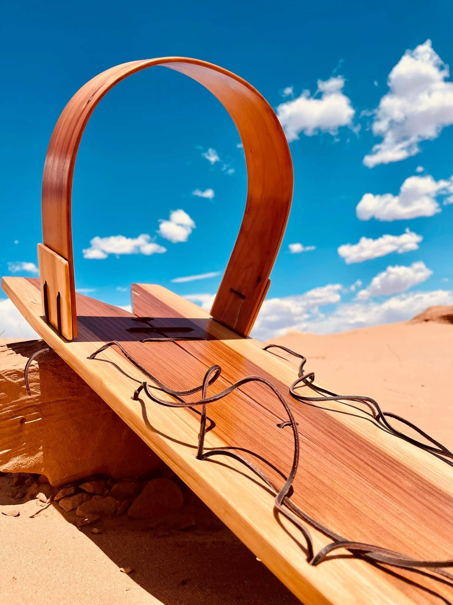 A wooden sled with a curved handle resting on sand under a partly cloudy blue sky.