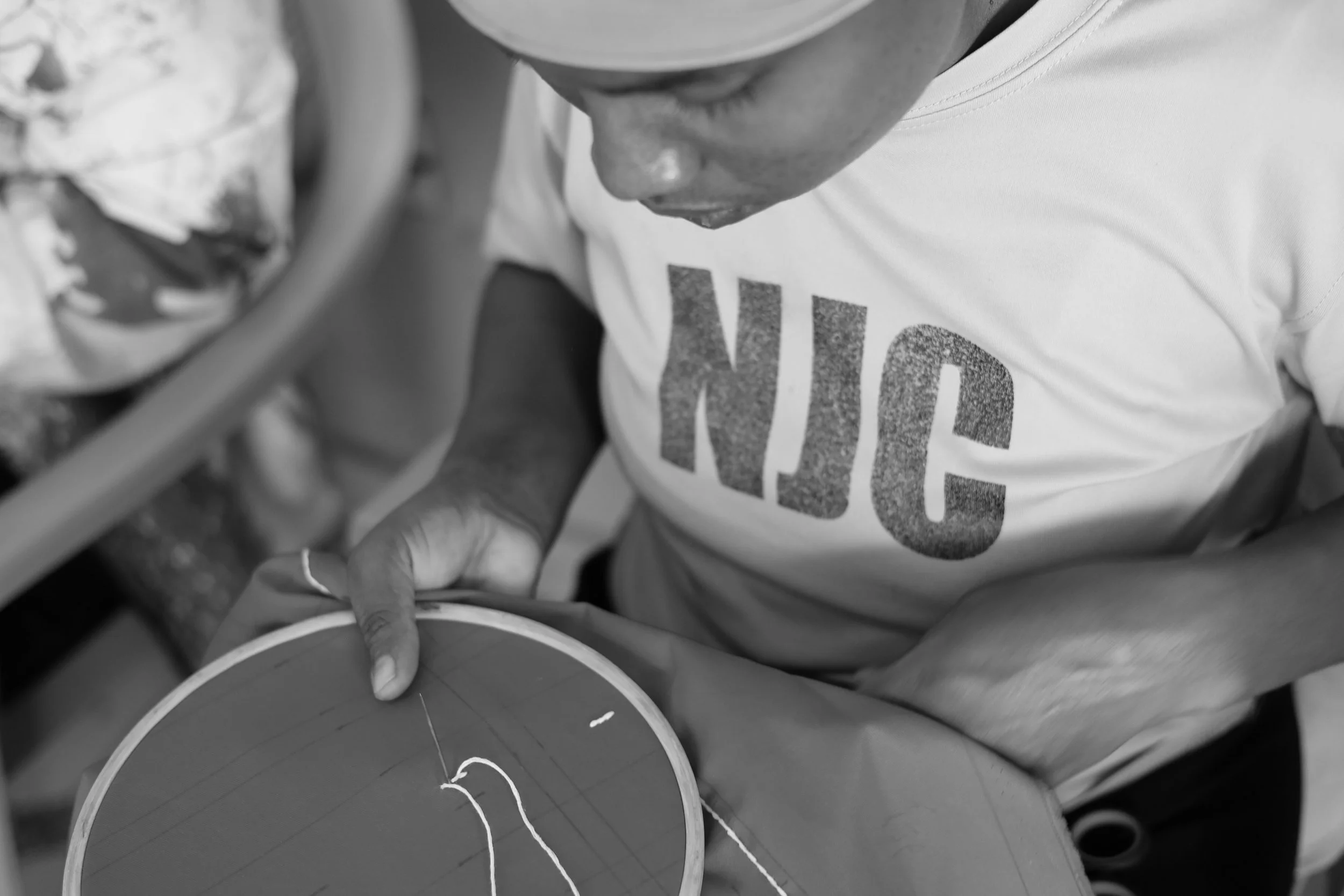 A person working on embroidery, holding a hoop with fabric and thread, wearing a t-shirt with the word 'NICE' on it, in black and white.