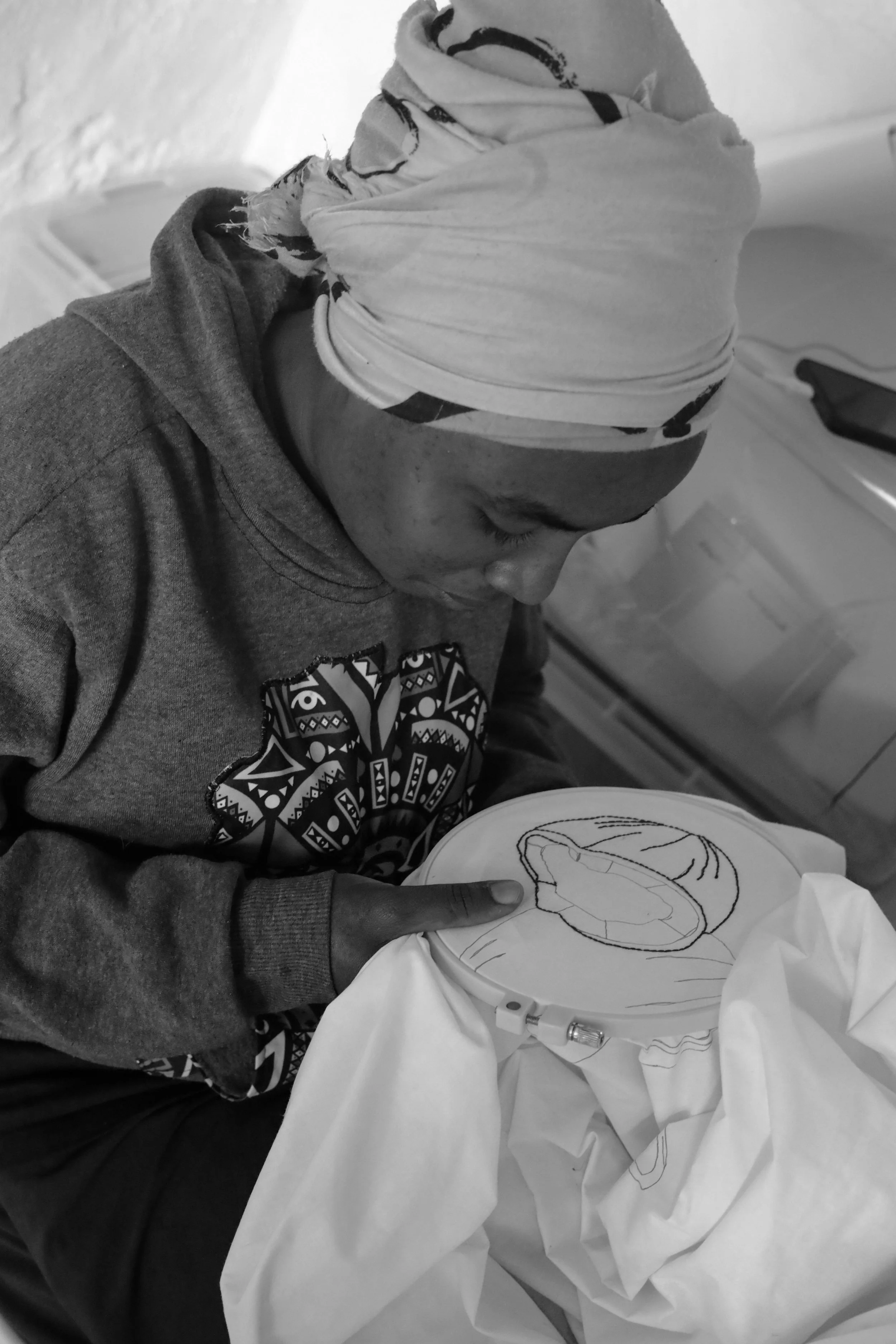 A young woman with a patterned headscarf looking down at embroidery hoop with fabric, working on a design.