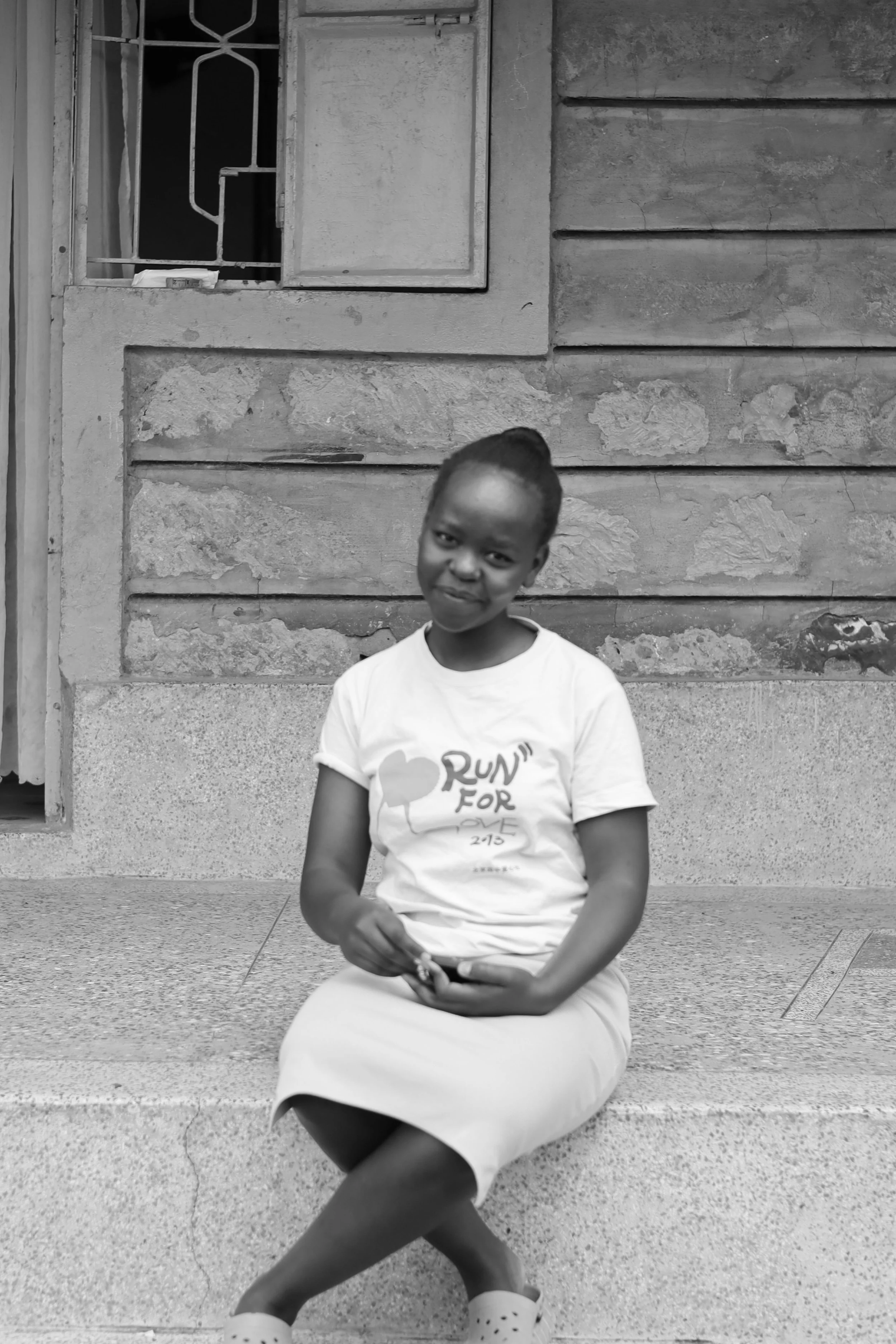 A young girl sitting on a concrete ledge outside a building, smiling slightly while holding a phone in her hand.