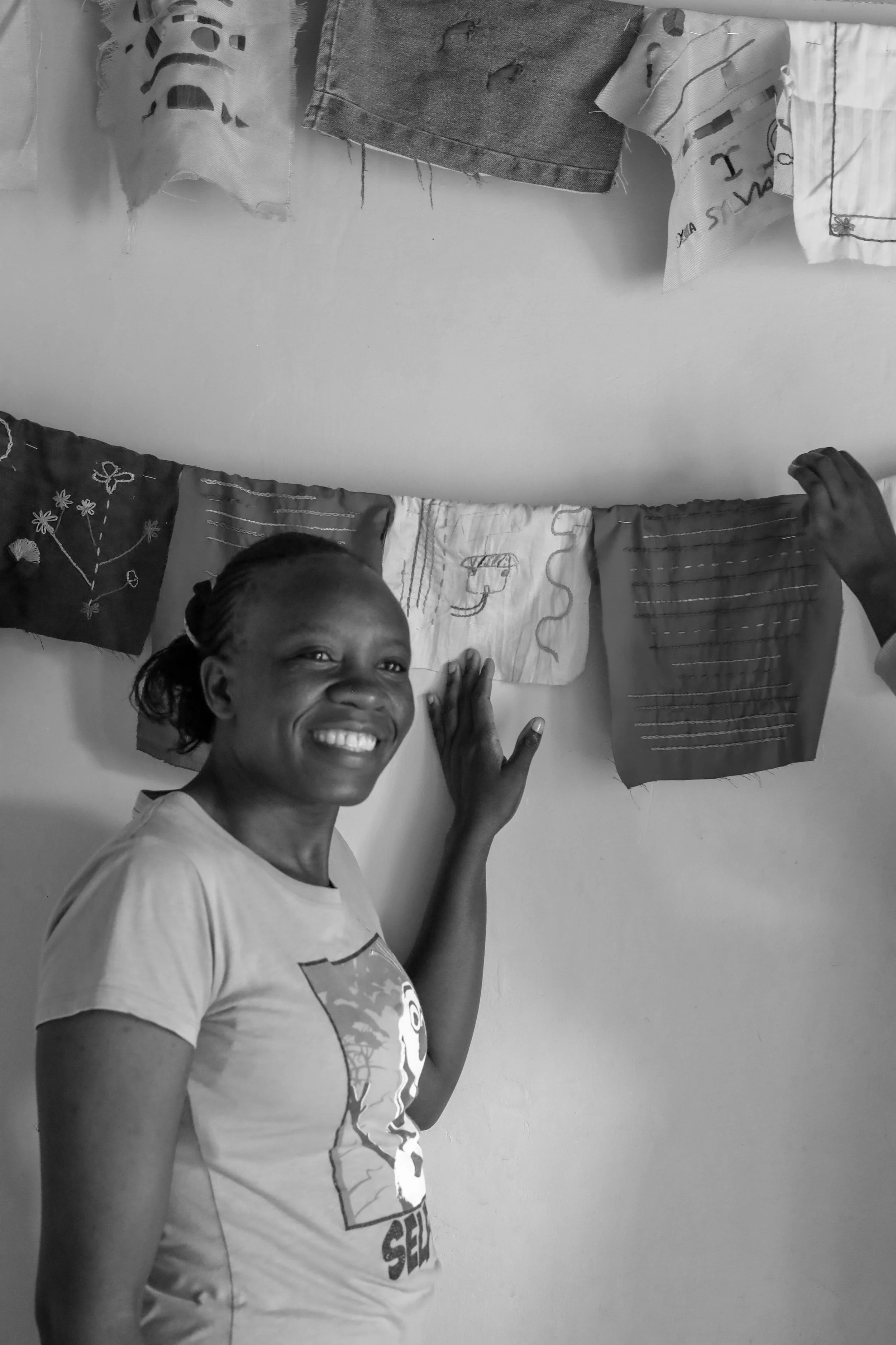 A woman with a bright smile, standing beside a wall of hanging pieces of fabric or clothing with embroidery and patterns.