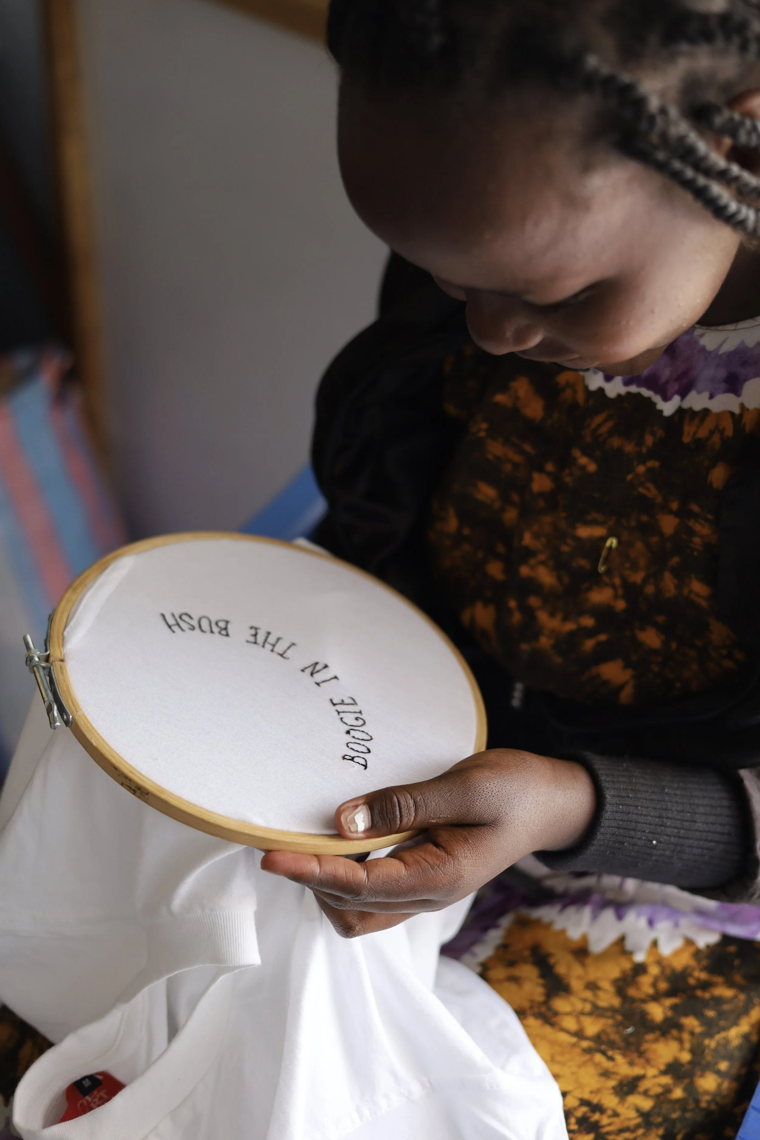 A person is looking down at an embroidery hoop with white fabric, which has the phrase 'BOGGLE IN THE BUSH' embroidered on it.
