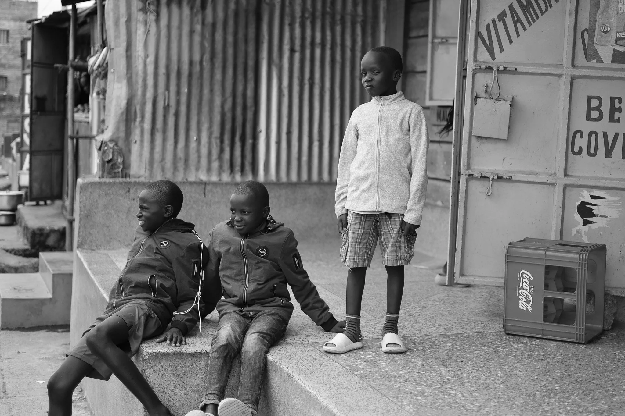 Three children sitting and standing on a sidewalk in an urban area. The two children sitting are smiling, while the child standing looks serious. The background includes corrugated metal walls, a Coca-Cola crate, and signage.