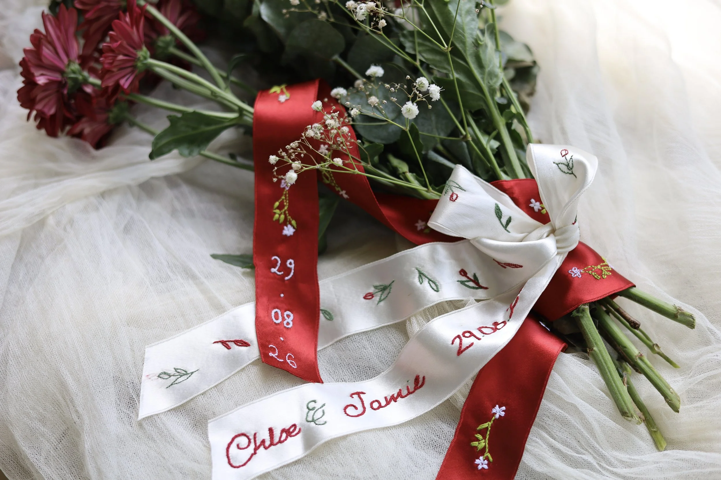 A bouquet of red and pink flowers with green leaves, wrapped with white, red, and white ribbons embroidered with floral designs and dates, resting on white fabric.