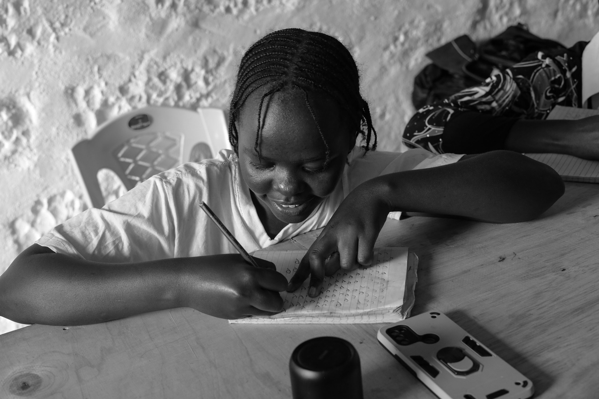 A young girl with braided hair is sitting at a wooden table, writing in a notebook with a pen. There is a smartphone on the table in front of her, and a backpack in the background. The background wall is made of textured stone.