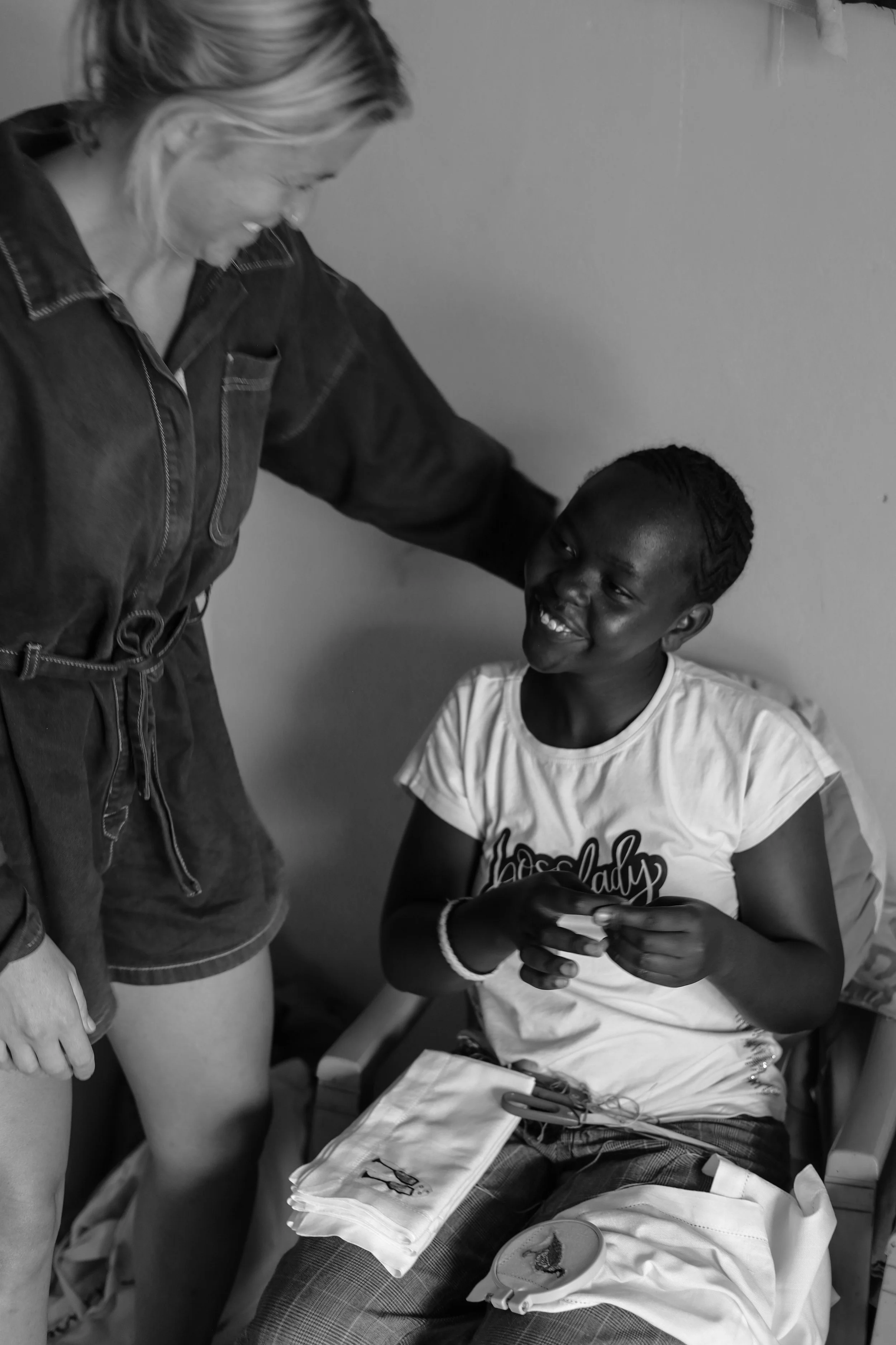 A smiling girl sitting on a bed, holding a small object, with a woman leaning over her and touching her head, in a room with a plain wall.