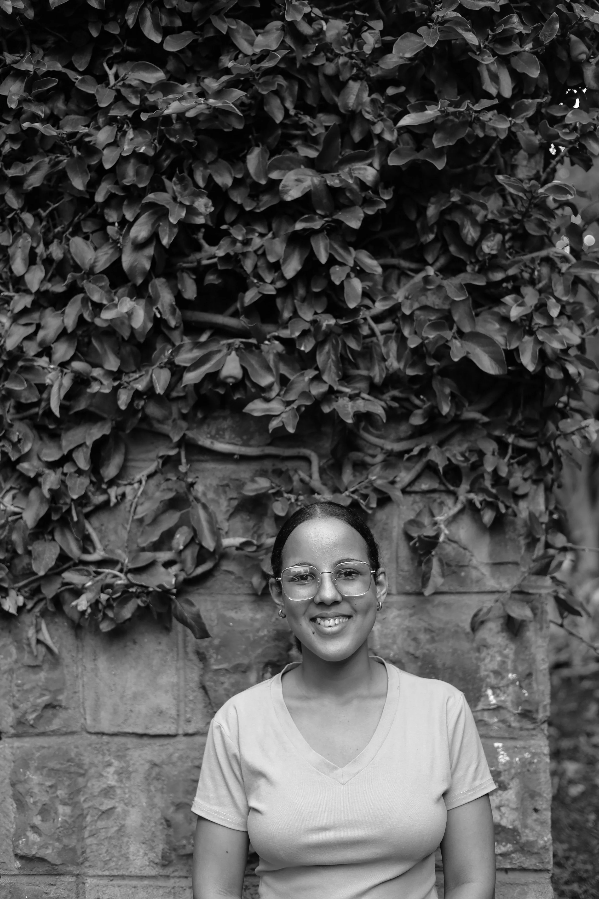A smiling woman wearing glasses and earrings standing outdoors in front of a stone wall covered with dense foliage.