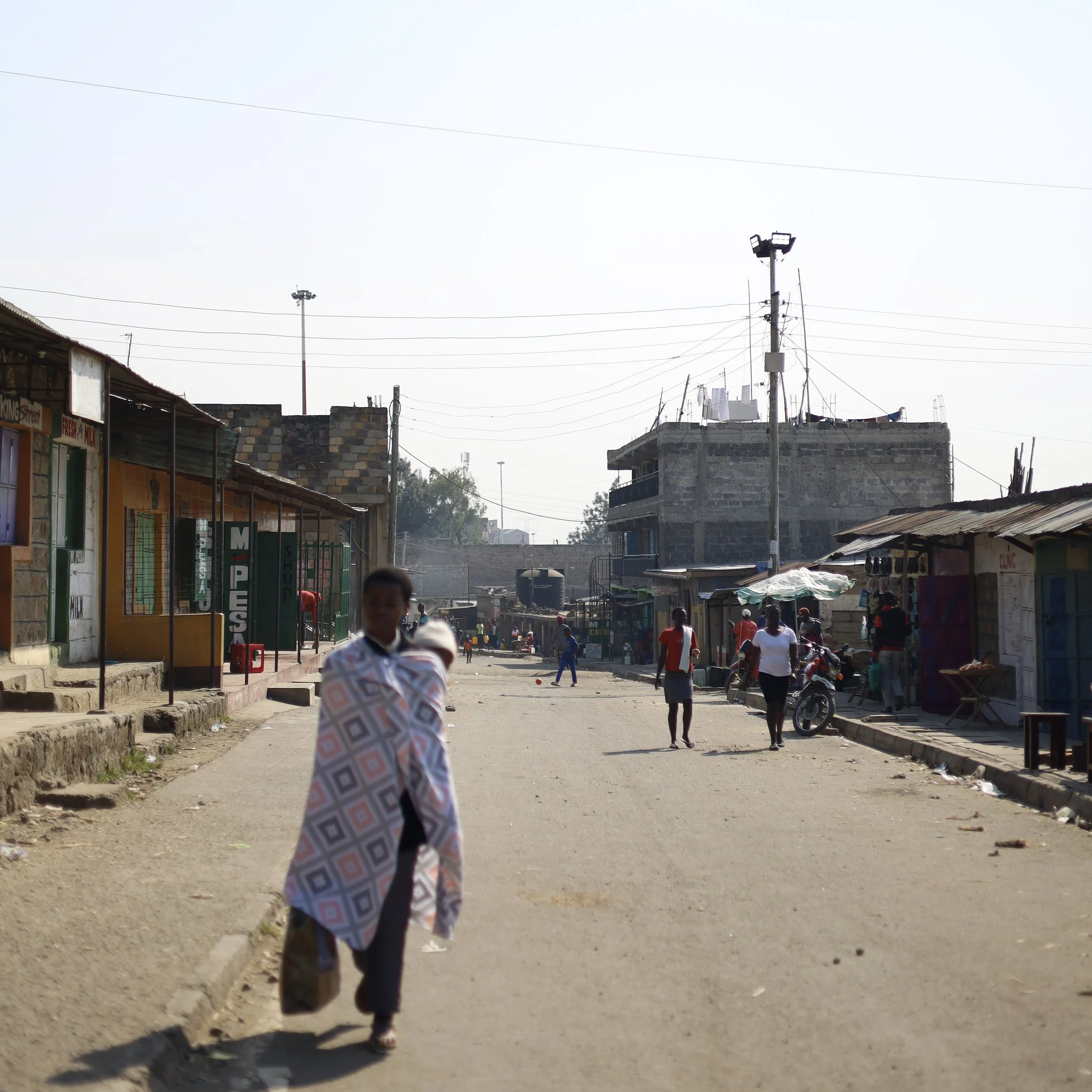 A street scene in a developing area with several people walking, some shops on the left side, and a large unfinished or under construction building in the background. The street is unpaved.