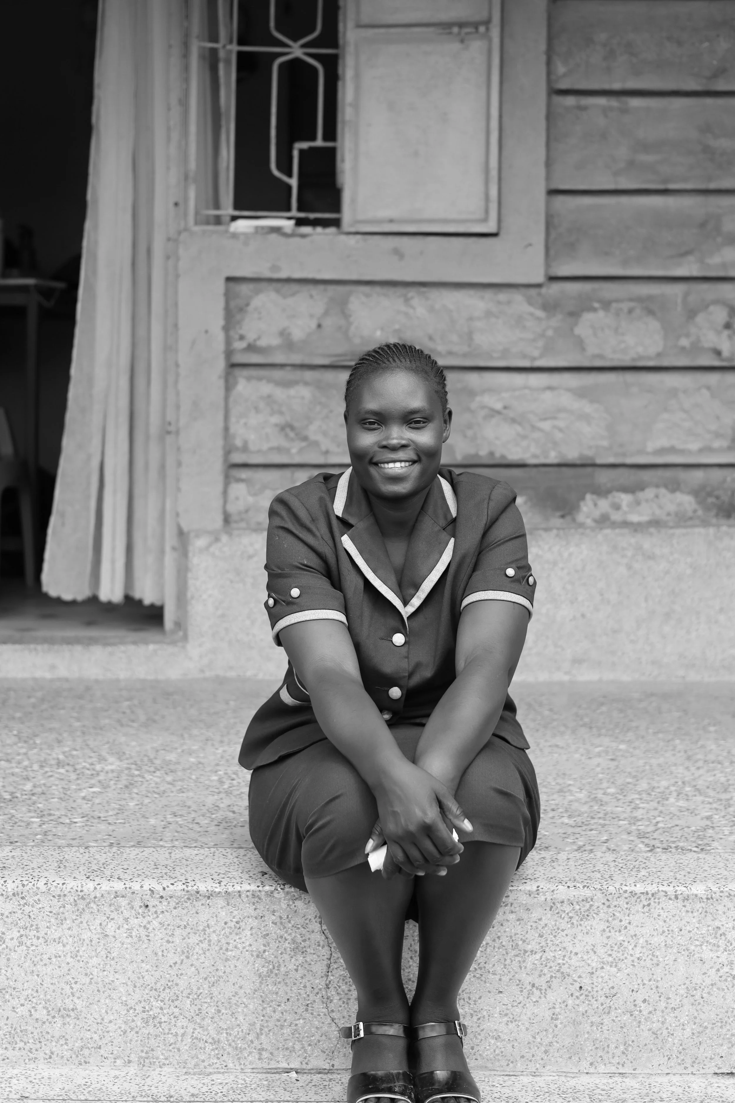 A young woman with braided hair, dressed in a dark uniform with light-colored trim and buttons, sitting on concrete steps outside a wooden house, smiling at the camera.