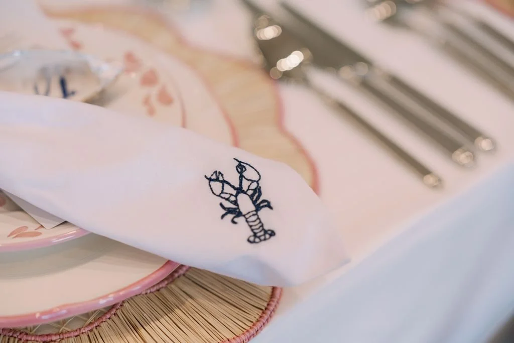 A white napkin with a lobster embroidery, placed on a pink plate with a floral design, partly covering a bamboo placemat.
