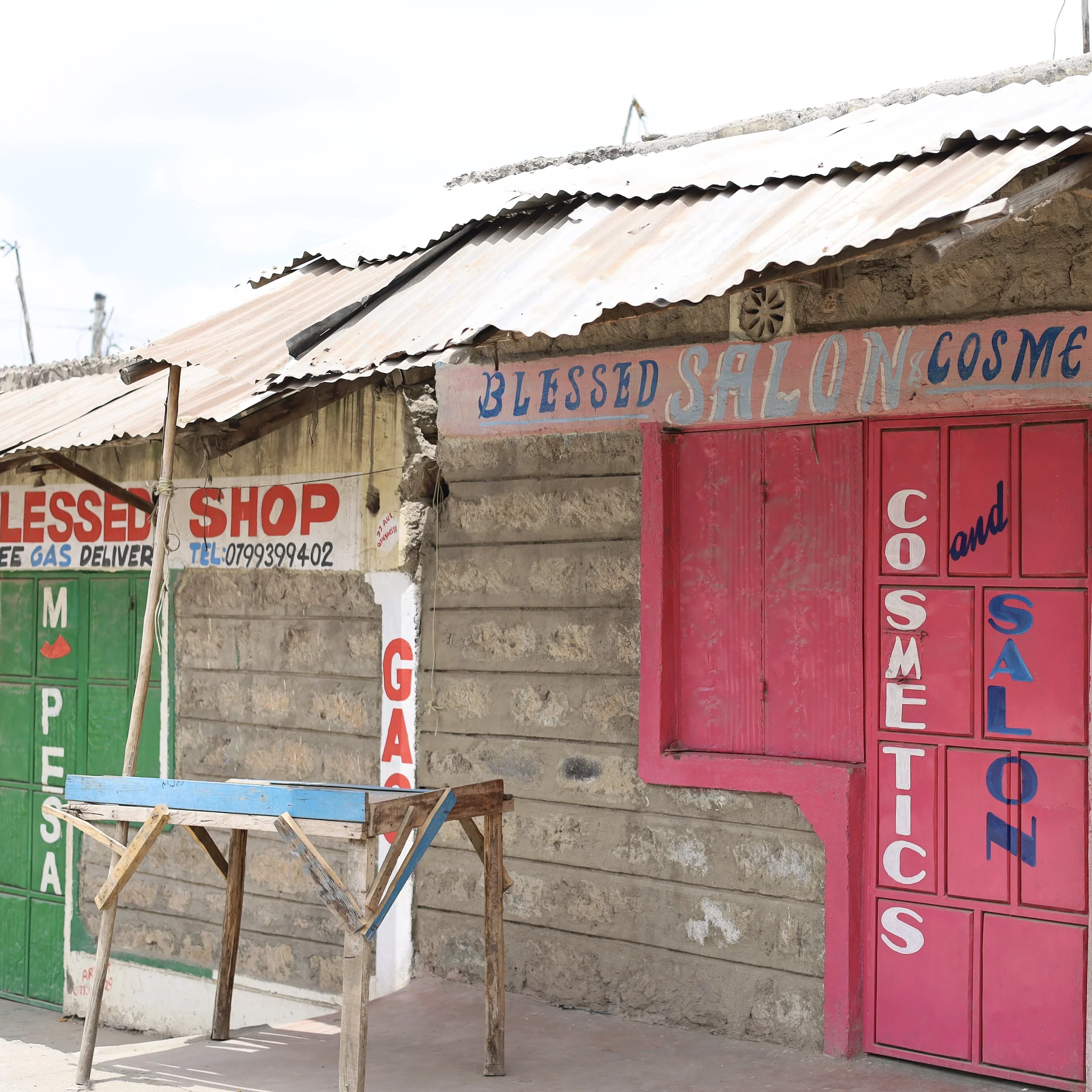 Exterior of a small shop with colorful painted signs reading 'Blessed Shop,' 'Gas,' 'Cosmetics,' and 'Saloon,' located in a rural area with a corrugated metal roof.