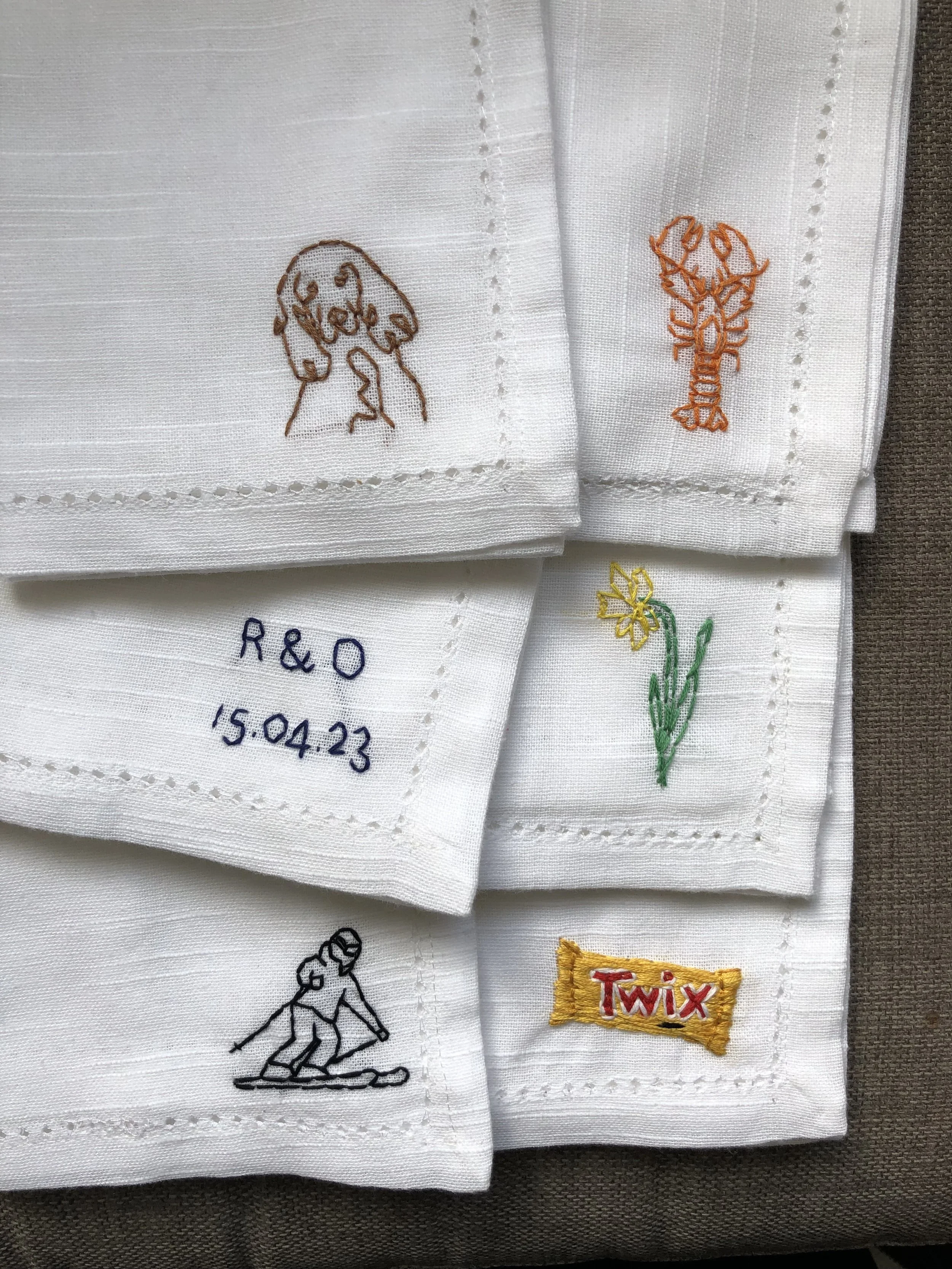 A white cloth with embroidered outlines of a dog, a lobster, a yellow flower, a skier, and the text 'R & O 15.04.23' and the Twix logo.
