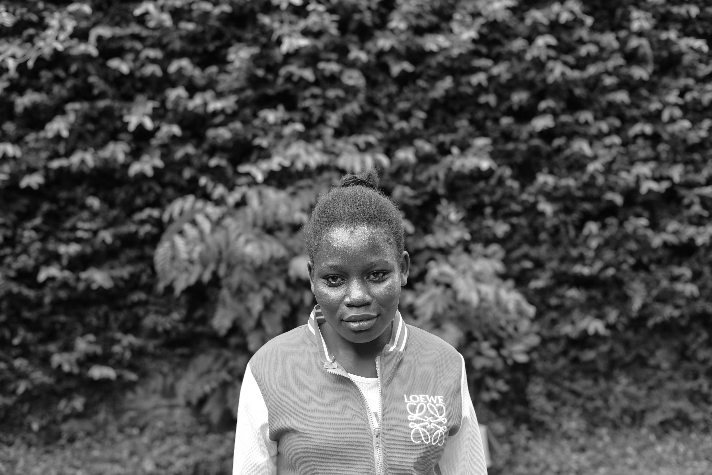 Black and white photo of a woman with a bun hairstyle wearing a jacket with the word "LOEWE" on it, standing outdoors in front of a background full of trees and foliage.