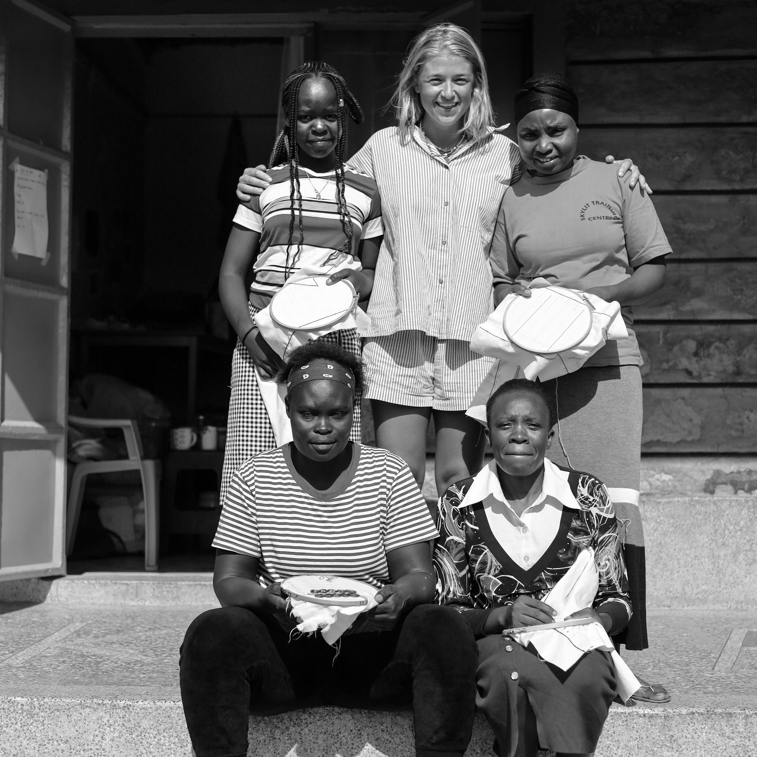 Group of six women and girls posing outdoors, some holding cloth, with two seated in front and four standing behind, smiling.