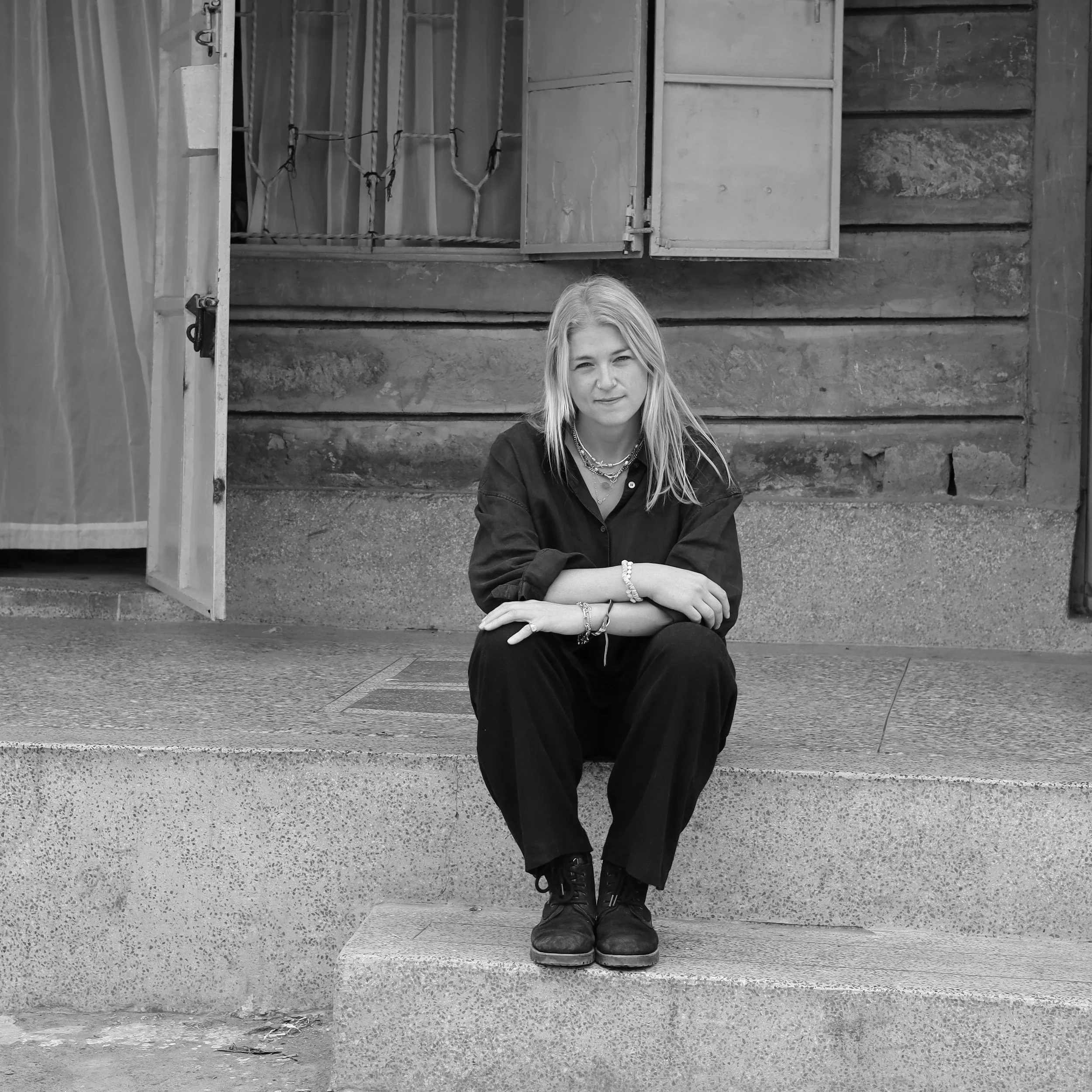 A young woman with long blonde hair sitting on a concrete step outside a rustic wooden building, wearing a dark shirt and pants with jewelry accessories.