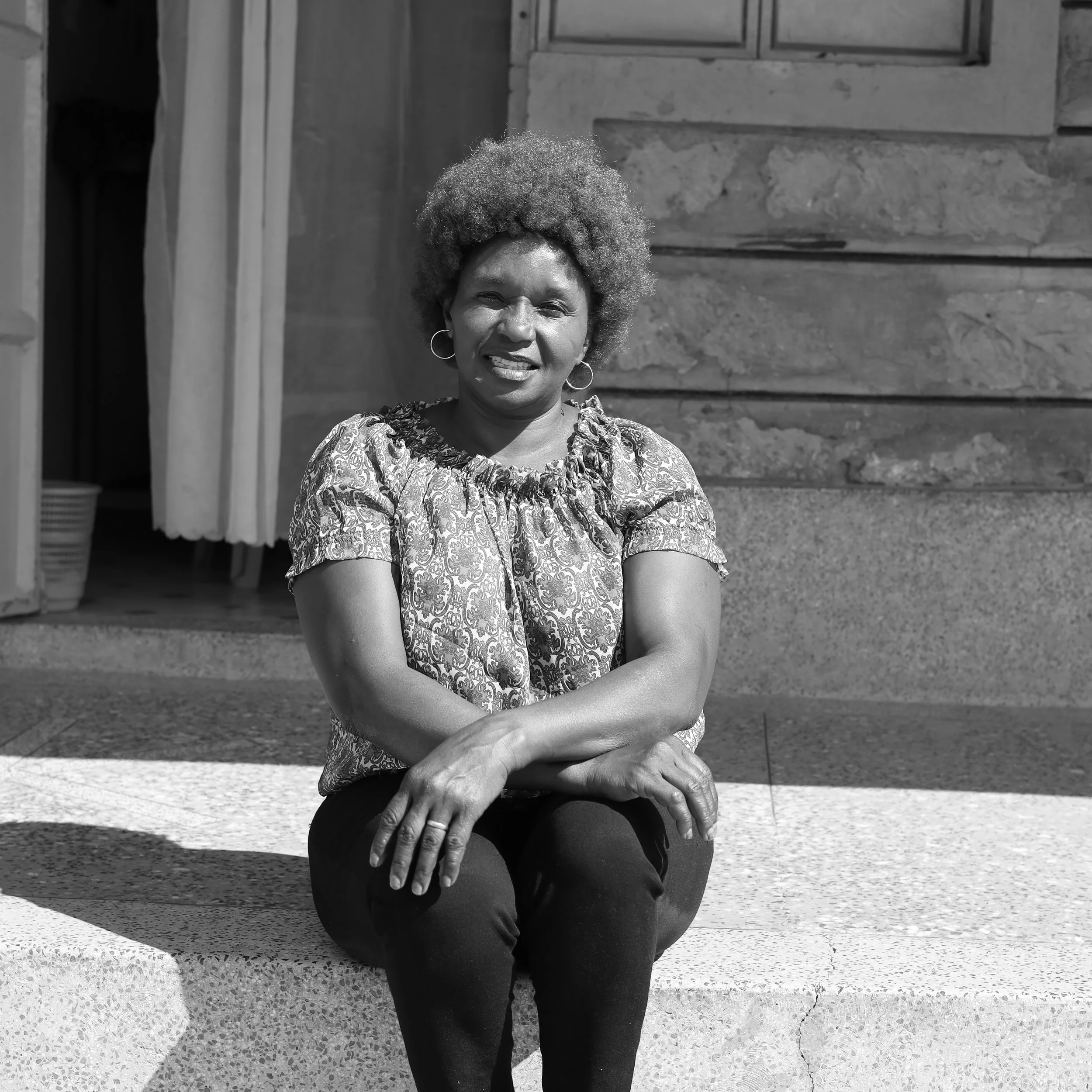 A woman sitting on a concrete step outside a building, smiling, with short curly hair, wearing hoop earrings and a patterned blouse.