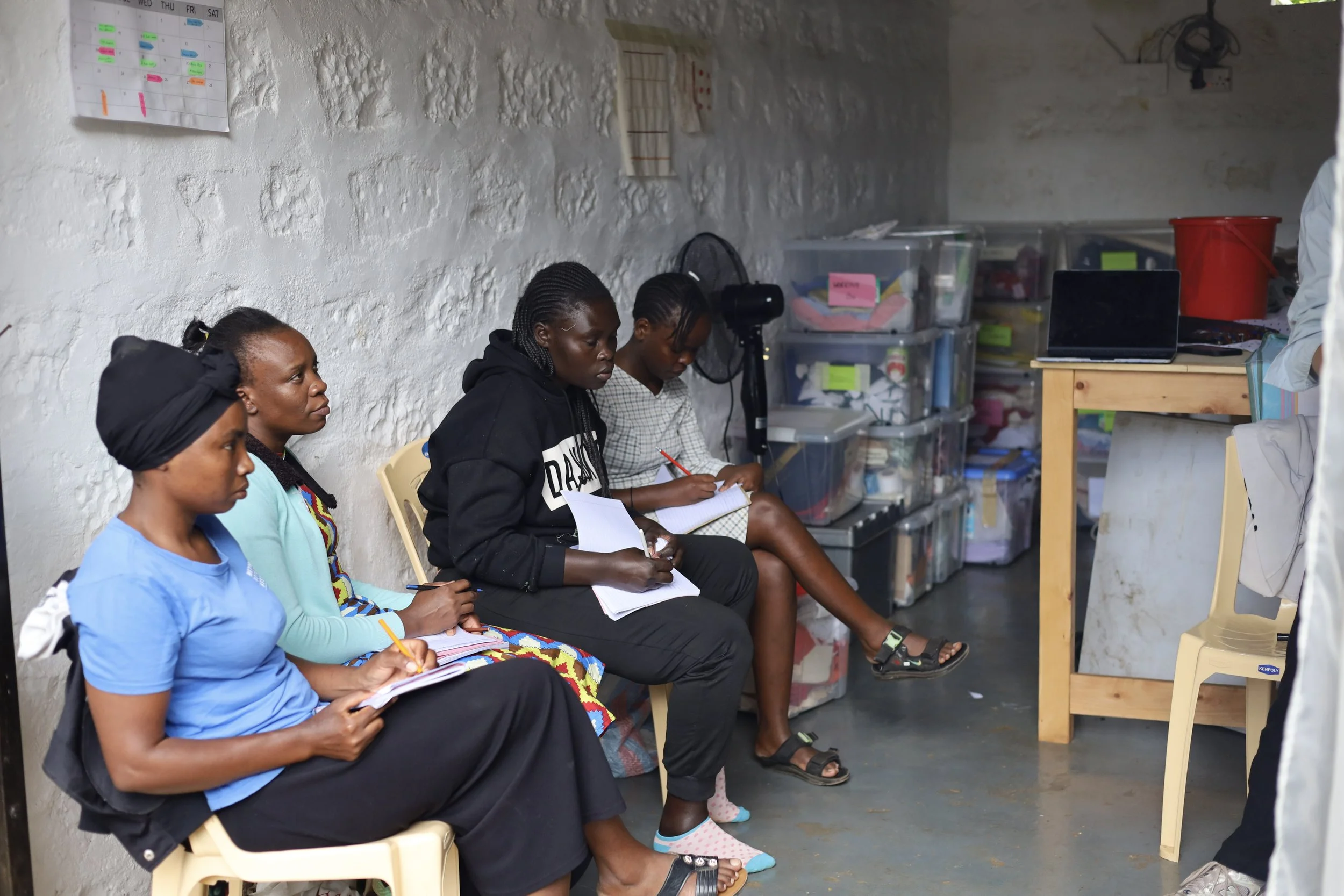 Four women sitting on chairs in a room, taking notes, with storage boxes and a desk with a laptop in the background.
