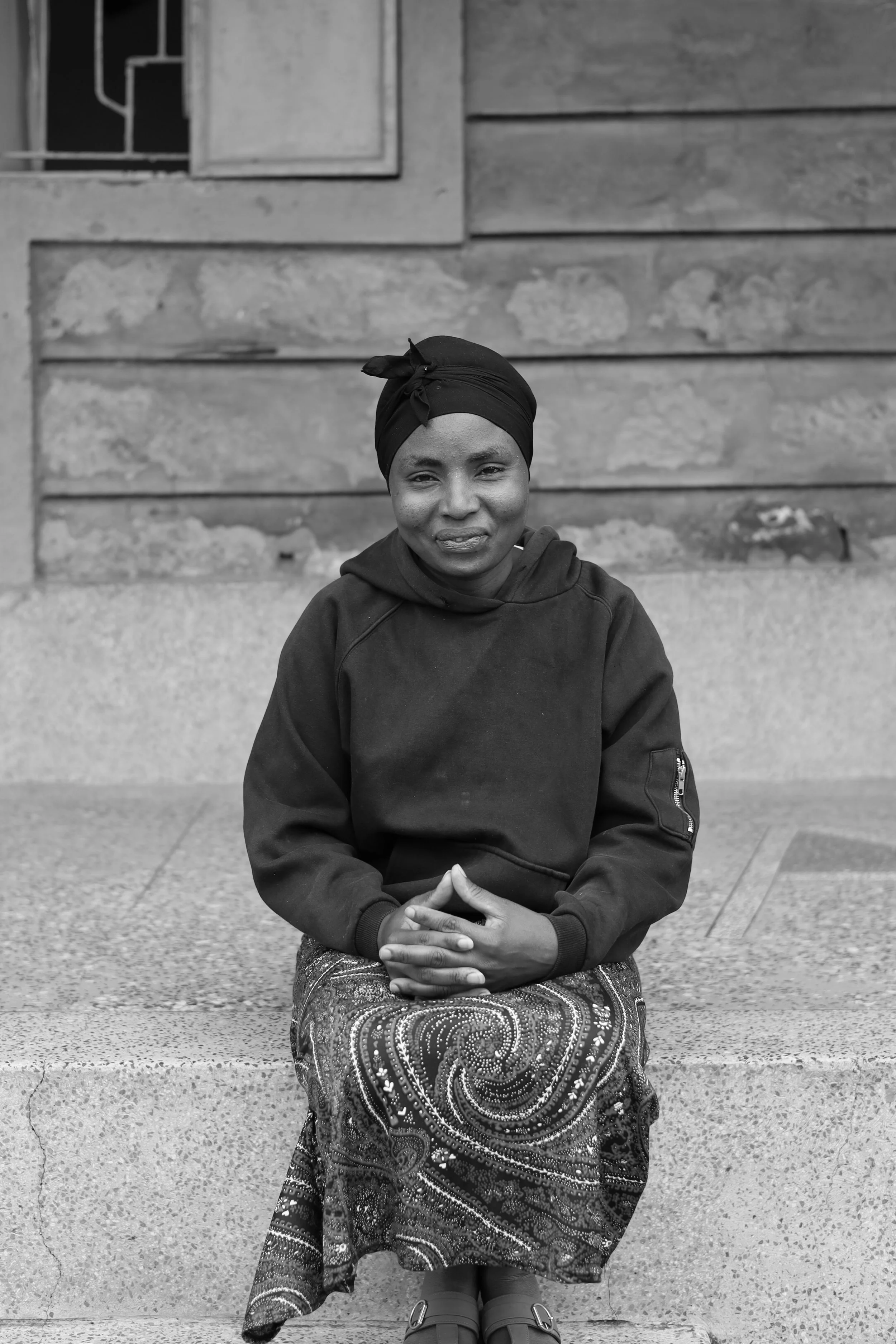 A woman sitting on a concrete step, wearing a headscarf and a patterned skirt, with a wooden building in the background.