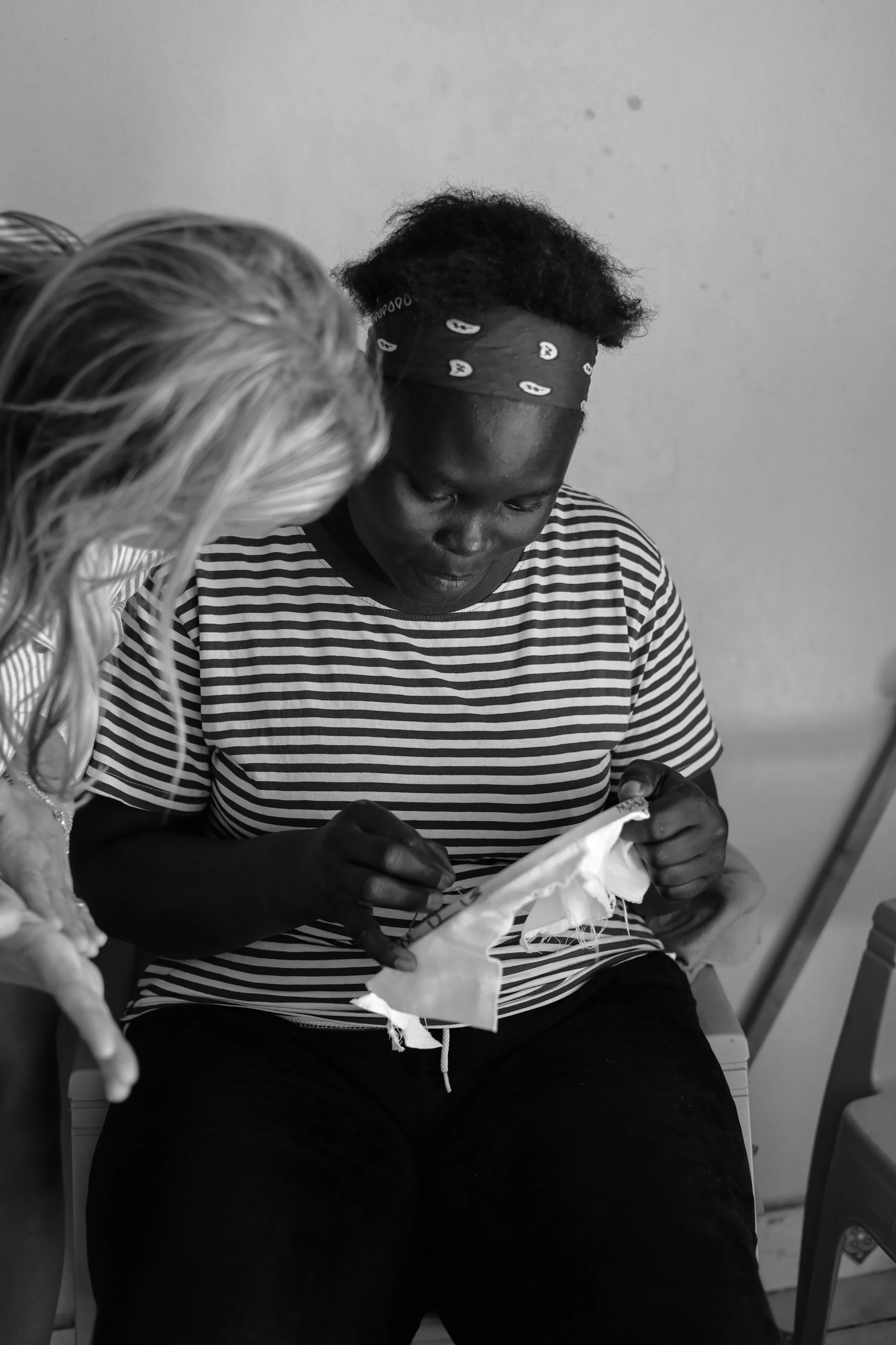 A woman with dark skin, wearing a striped shirt and a bandana, sitting in a room, looking at and holding a piece of cloth in her hands while a blonde person leans in to look at the cloth.