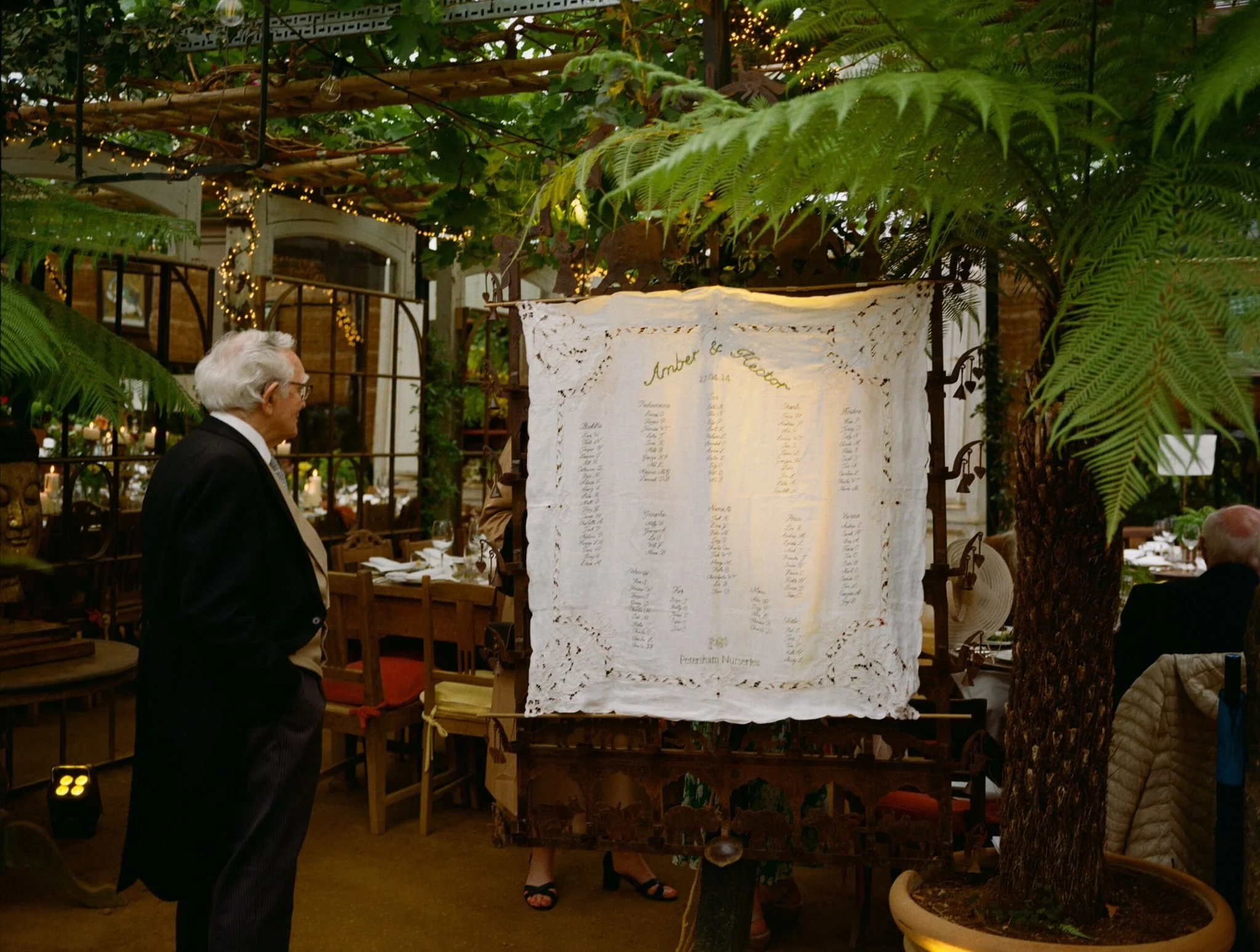 An elderly man with white hair, glasses, wearing a black tuxedo, standing in a decorated greenhouse or conservatory with green plants and fairy lights. He is looking at a large, lace-edged seating chart or program on display.