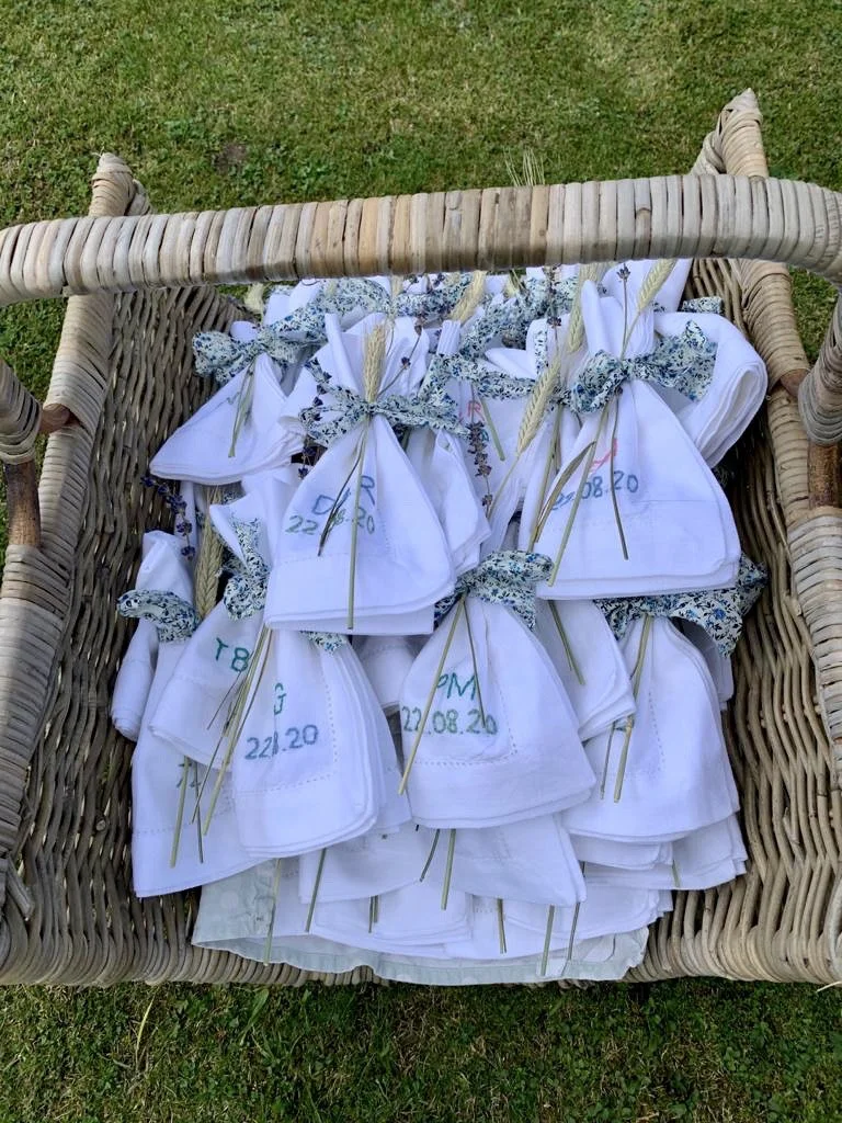 A wicker basket filled with small white cloth bags tied with blue and green floral scarves and decorated with wheat stalks, set on grass. The bags are labeled with handwritten notes.