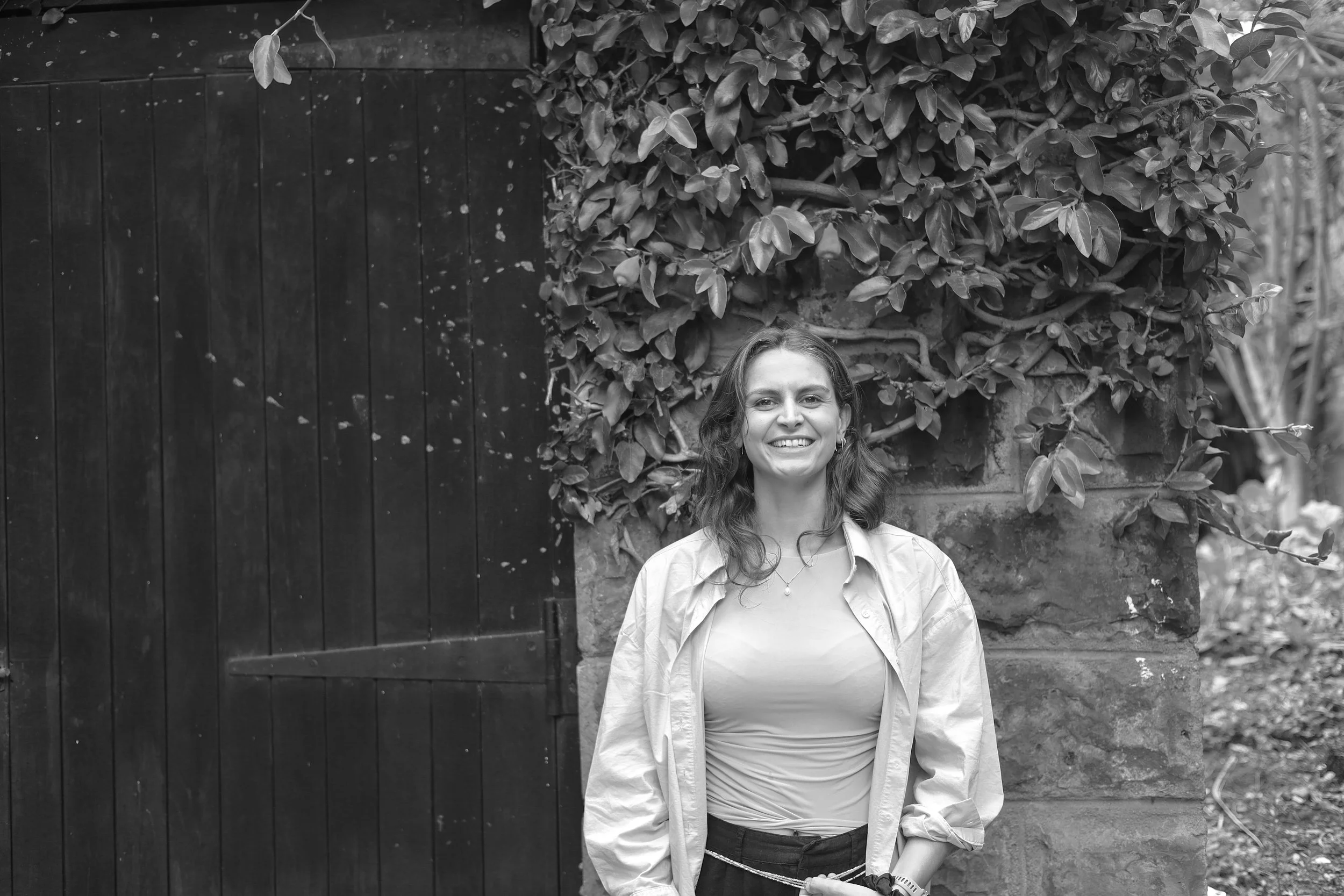 A woman smiling and standing outdoors in front of a brick wall with climbing plants, next to a dark wooden gate.