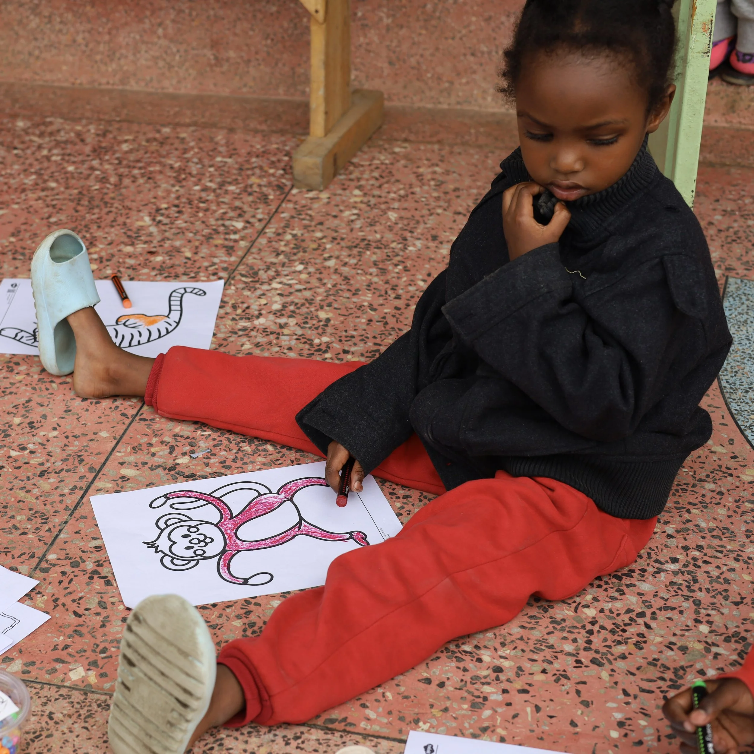 A young girl sitting on the floor coloring pictures of animals, with some coloring supplies and drawings around her.