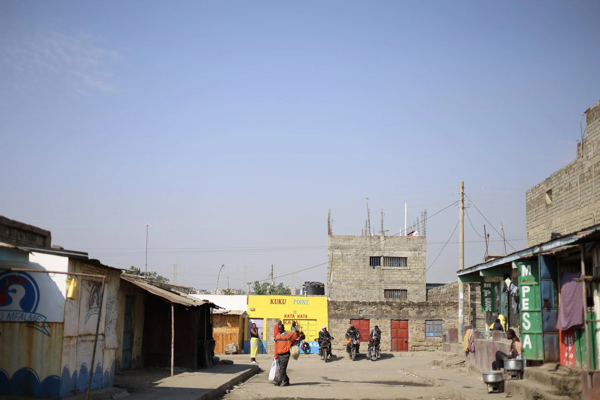 Street scene in a low-income area with small shops, motorcycles, and people walking under a clear blue sky.