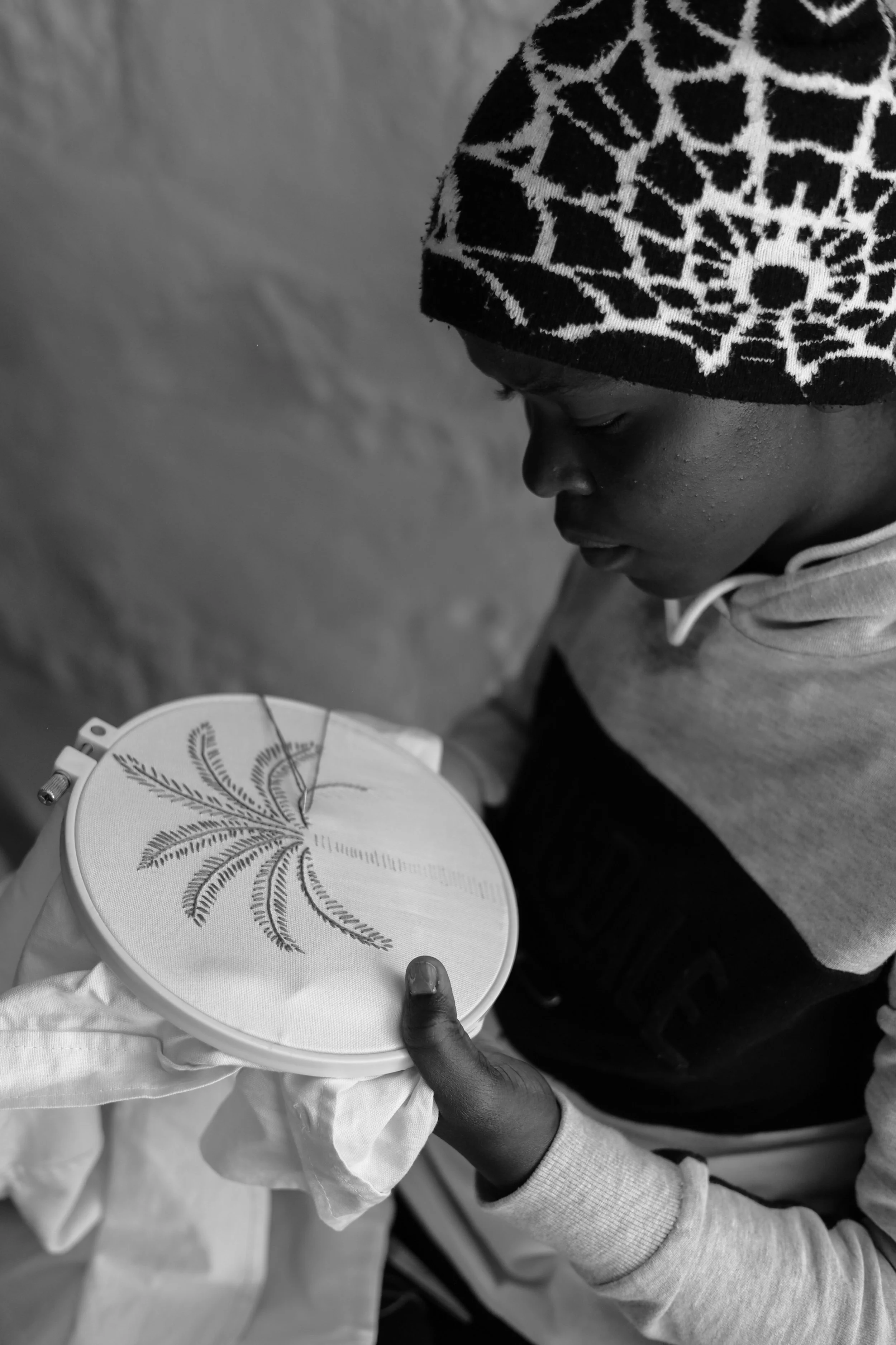 A young boy in a patterned beanie and hoodie, concentrating on stitching a leaf design on fabric in an embroidery hoop.