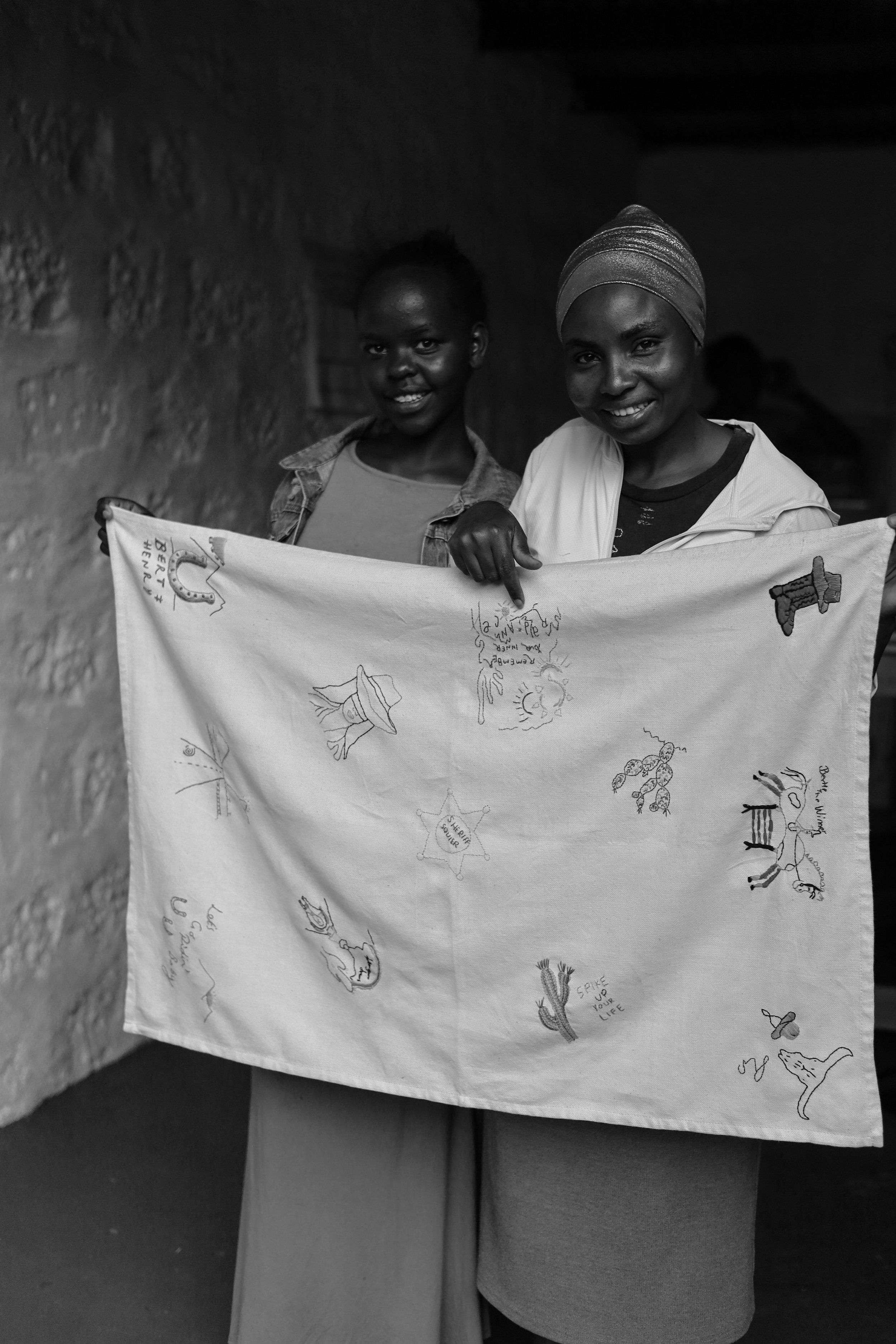 Two women smiling, holding up a decorated cloth with various drawings and writings. One woman is wearing a headscarf, and the other has short hair. They stand indoors against a textured wall.