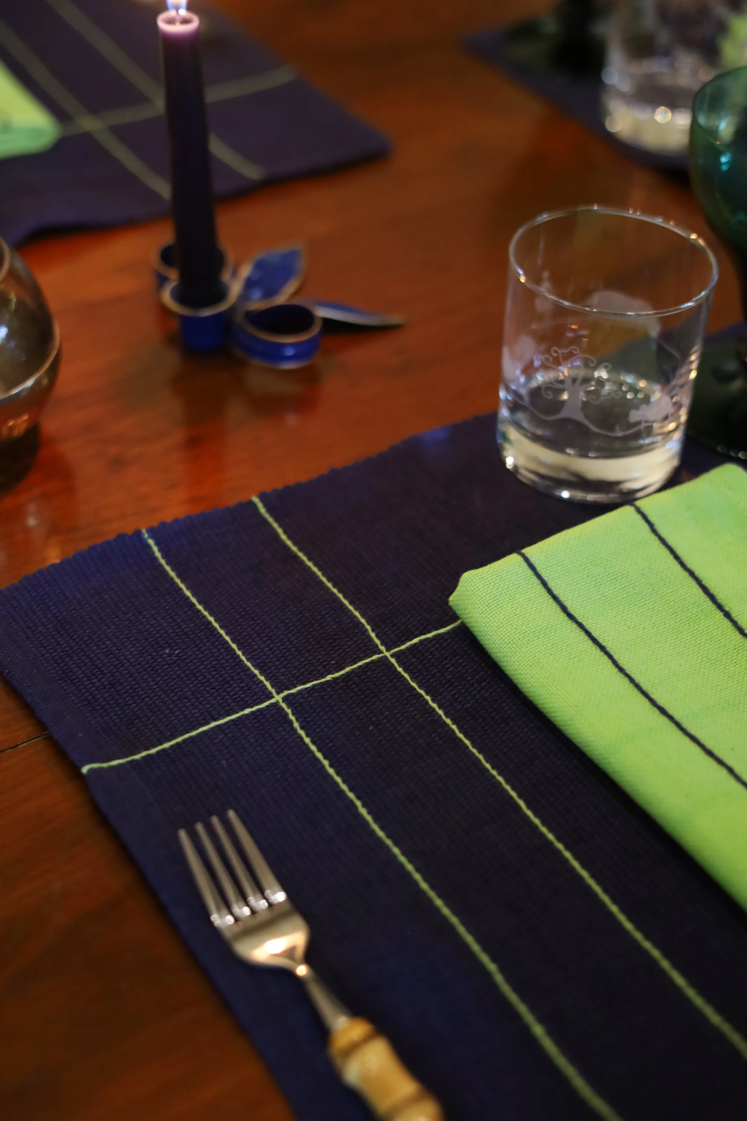Close-up of a dining table setting with a navy blue placemat, green napkin, silver fork, glass of water, and a lit purple candle in the background.